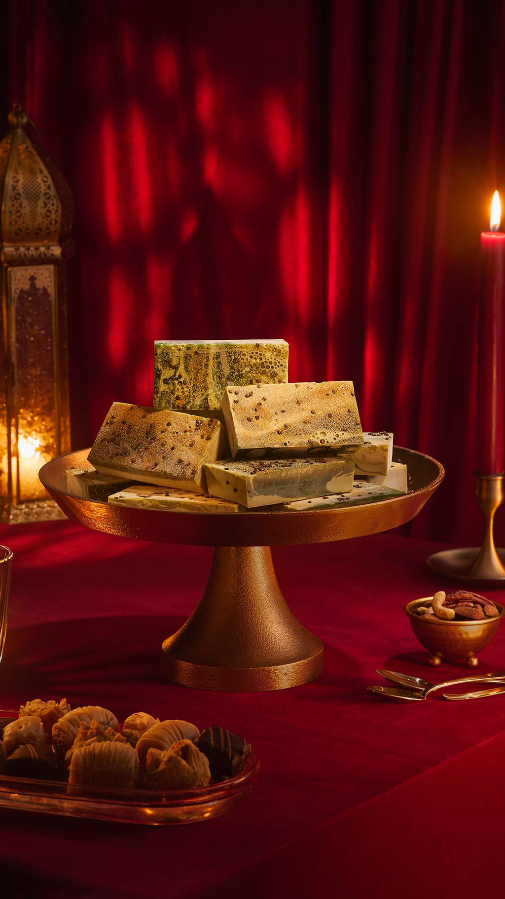 A stack of marbled green "Baklava" soap bars on a brass cake stand. The festive scene includes a red velvet tablecloth, a lit candle, an ornate gold lantern, and plates of baklava and nuts against a backdrop of draped red fabric.