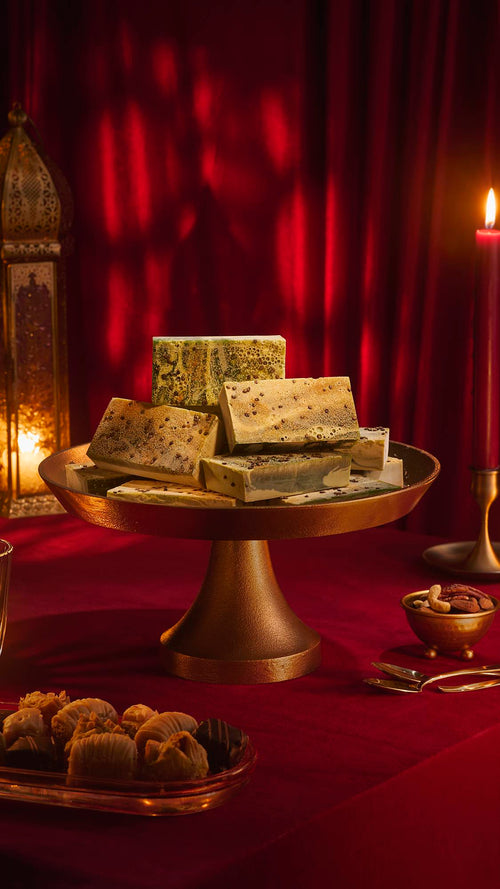 A stack of marbled green "Baklava" soap bars on a brass cake stand. The festive scene includes a red velvet tablecloth, a lit candle, an ornate gold lantern, and plates of baklava and nuts against a backdrop of draped red fabric.