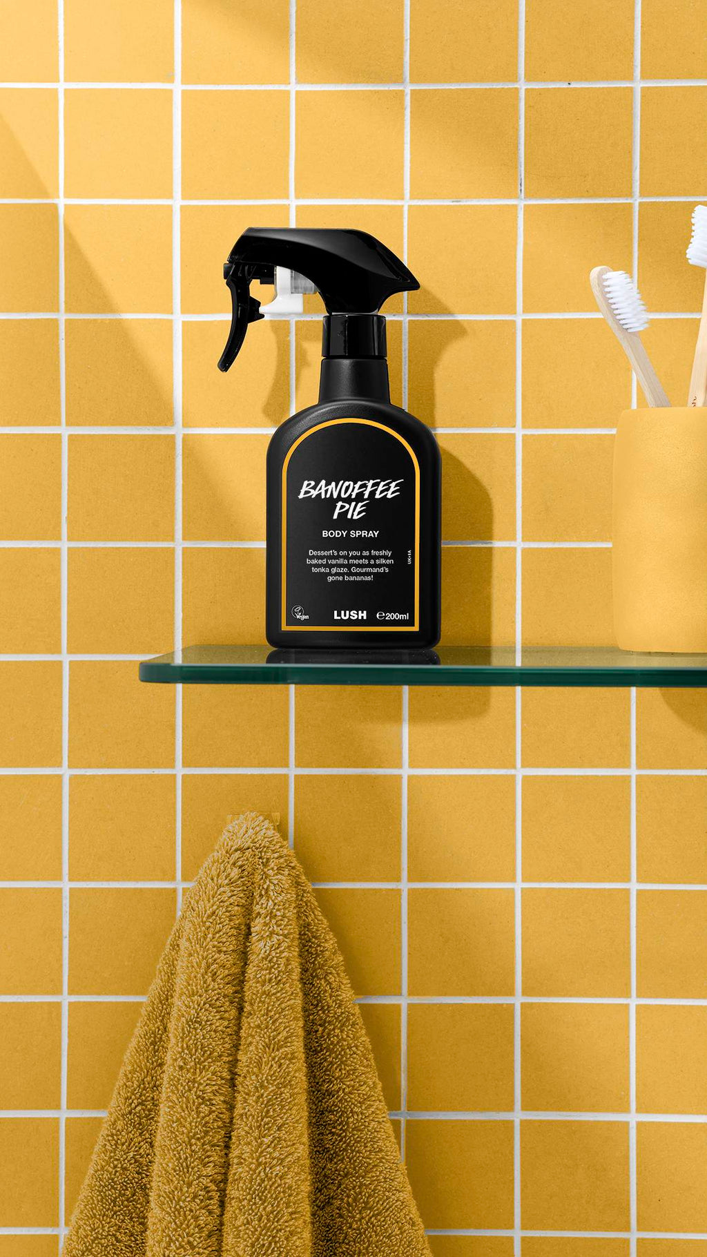 A black bottle of Lush 'Banoffee Pie' body spray sits on a glass shelf against a yellow tiled wall. To the right is a yellow cup holding toothbrushes, and below the shelf hangs a mustard-yellow towel.