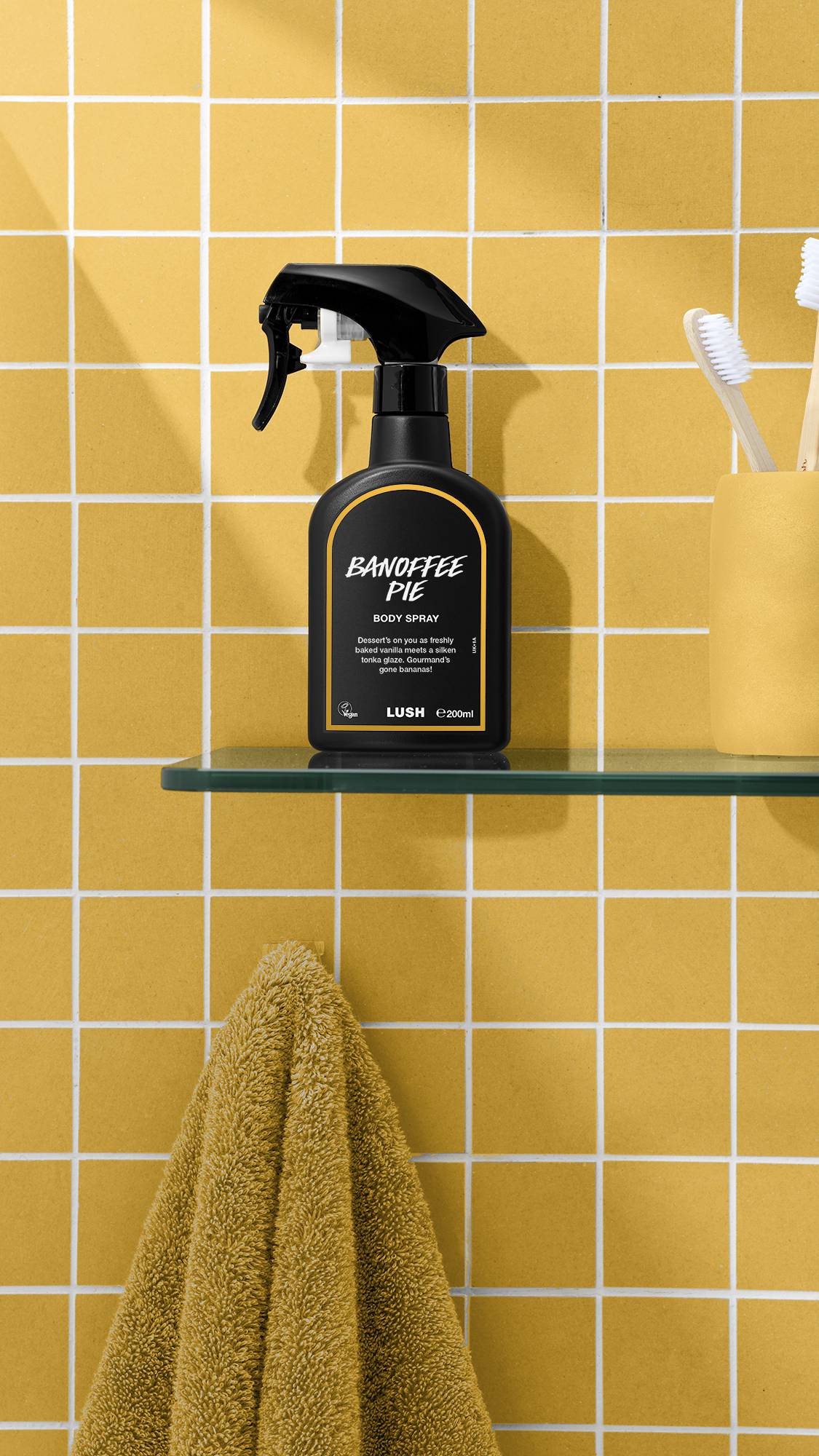 A black bottle of Lush 'Banoffee Pie' body spray sits on a glass shelf against a yellow tiled wall. To the right is a yellow cup holding toothbrushes, and below the shelf hangs a mustard-yellow towel.