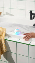 A round, white bath bomb-shaped waterproof speaker with glowing blue and rainbow lights sits on the edge of a white tiled bathtub filled with swirling bathwater. A hand rests on the tub nearby, next to a yellow striped towel.