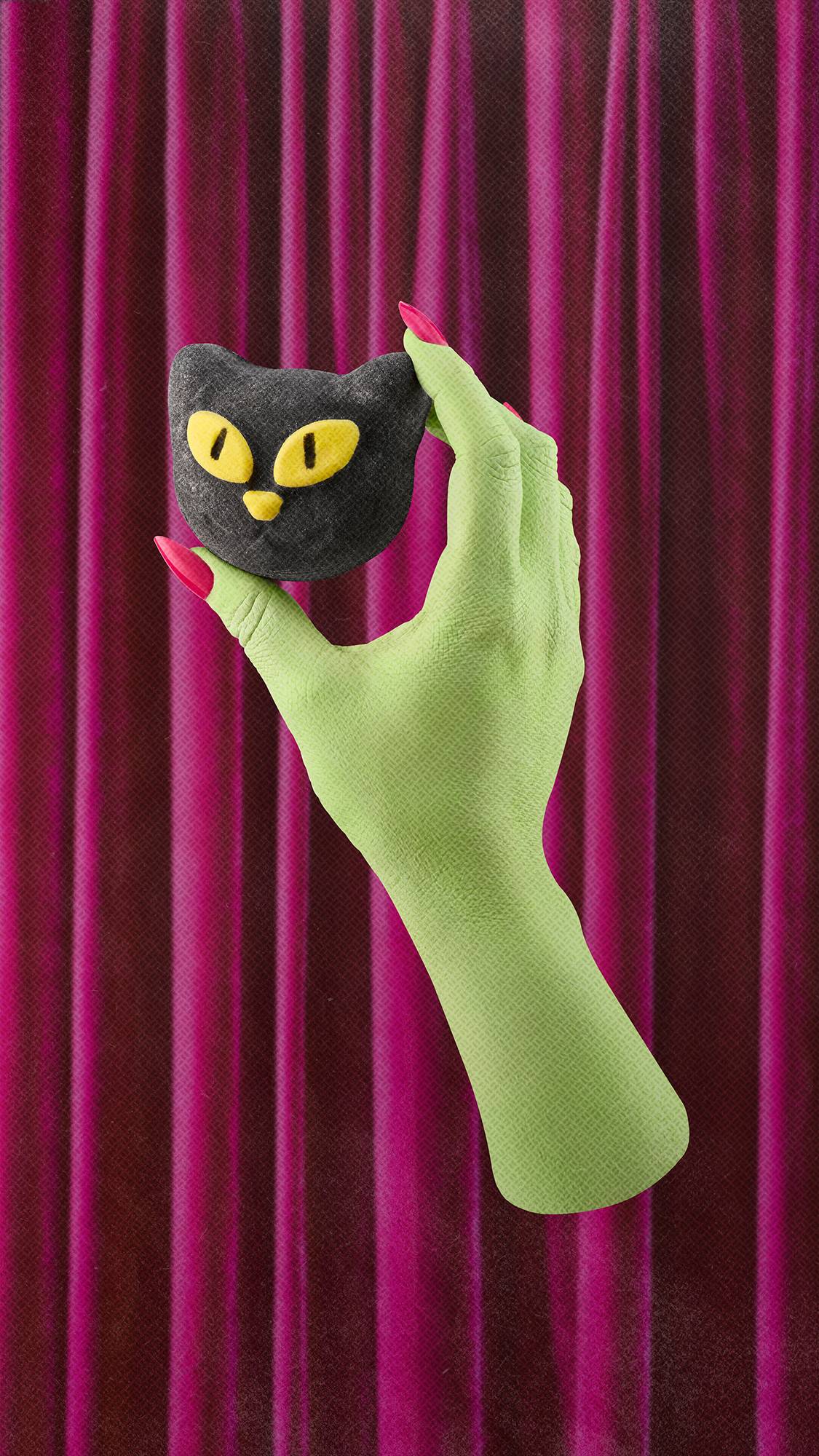 A lime green witch's hand with bright pink fingernails holds a black cat-shaped bubble bar with large yellow eyes. The hand is positioned against a background of draped magenta velvet curtains.