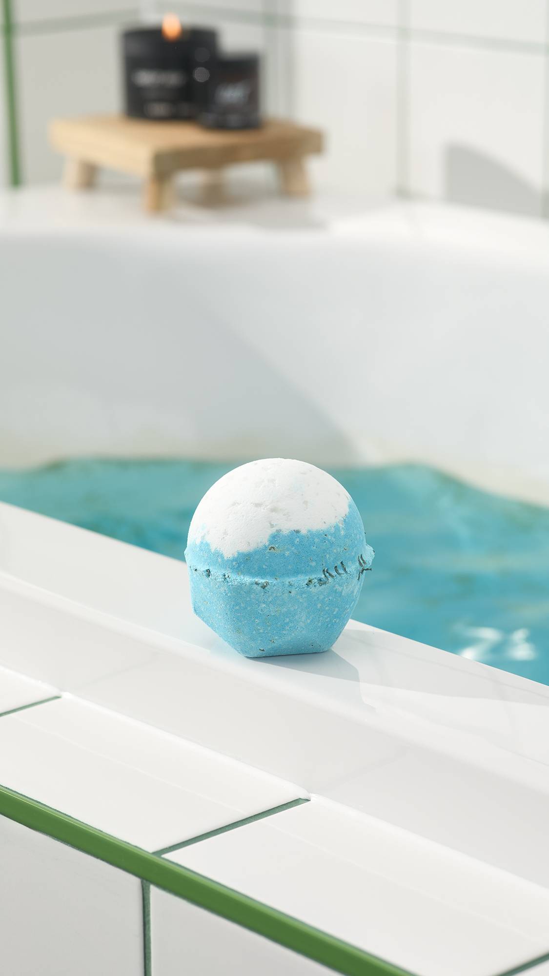 The blue and white Big Blue bath bomb sits on the white edge of a bathtub filled with bright blue water. In the background, a lit candle rests on a wooden stand against white tiles.