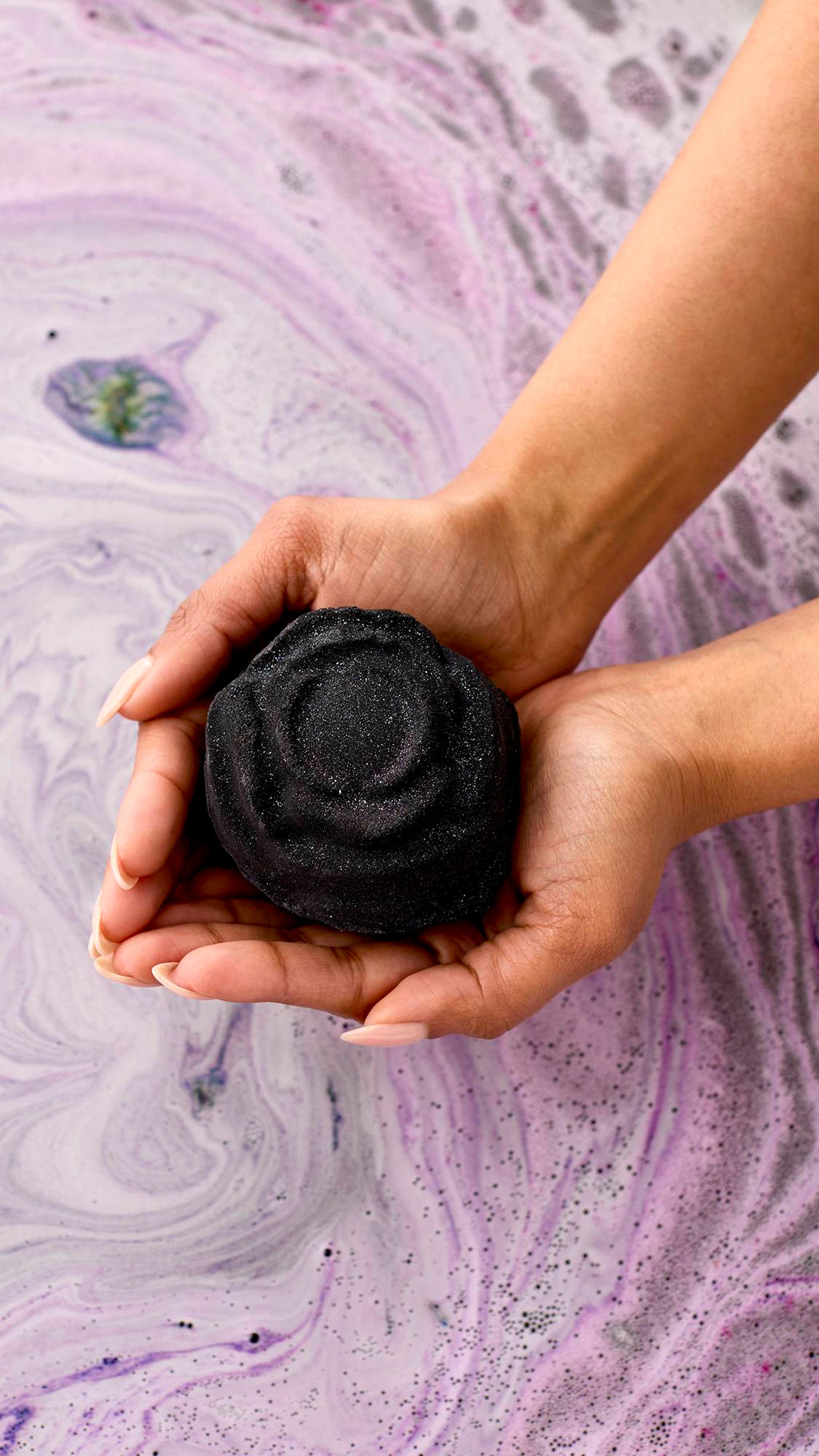 Black Rose. A dark, charcoal-grey bath bomb shaped like a blooming rose. It fizzes on the water's surface, releasing deep purple and pink swirls with a shimmering layer of silver plastic-free glitter.