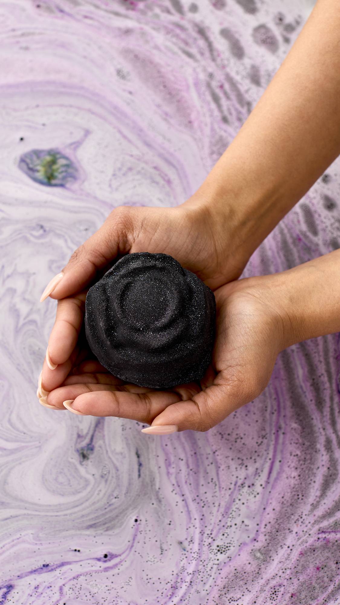 Black Rose. A dark, charcoal-grey bath bomb shaped like a blooming rose. It fizzes on the water's surface, releasing deep purple and pink swirls with a shimmering layer of silver plastic-free glitter.