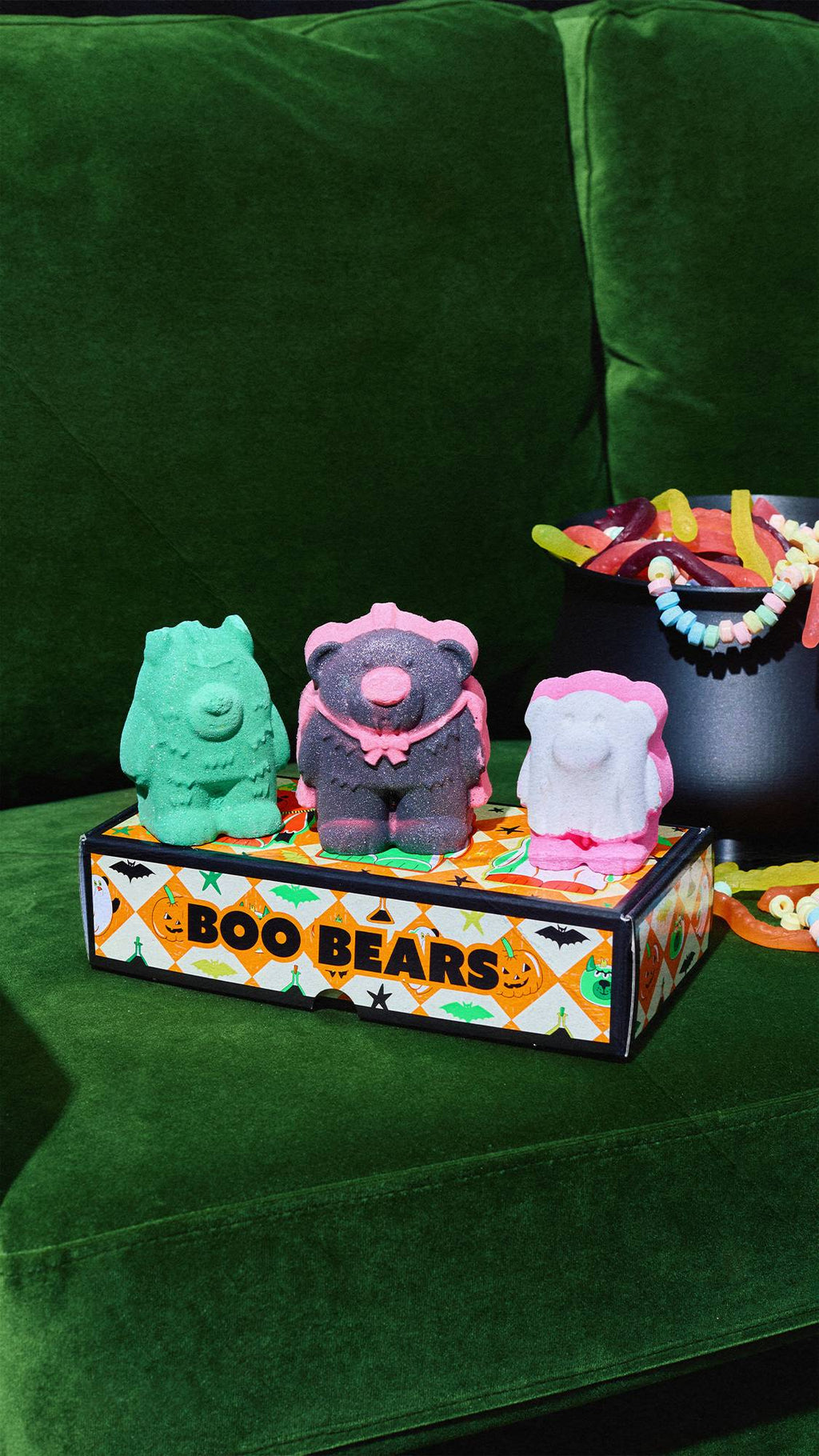 Three 'Boo Bears' monster bath bombs—green, vampire black, and white-and-pink—stand on their illustrated gift box on a green velvet couch. A cauldron filled with colourful sweets sits in the background.