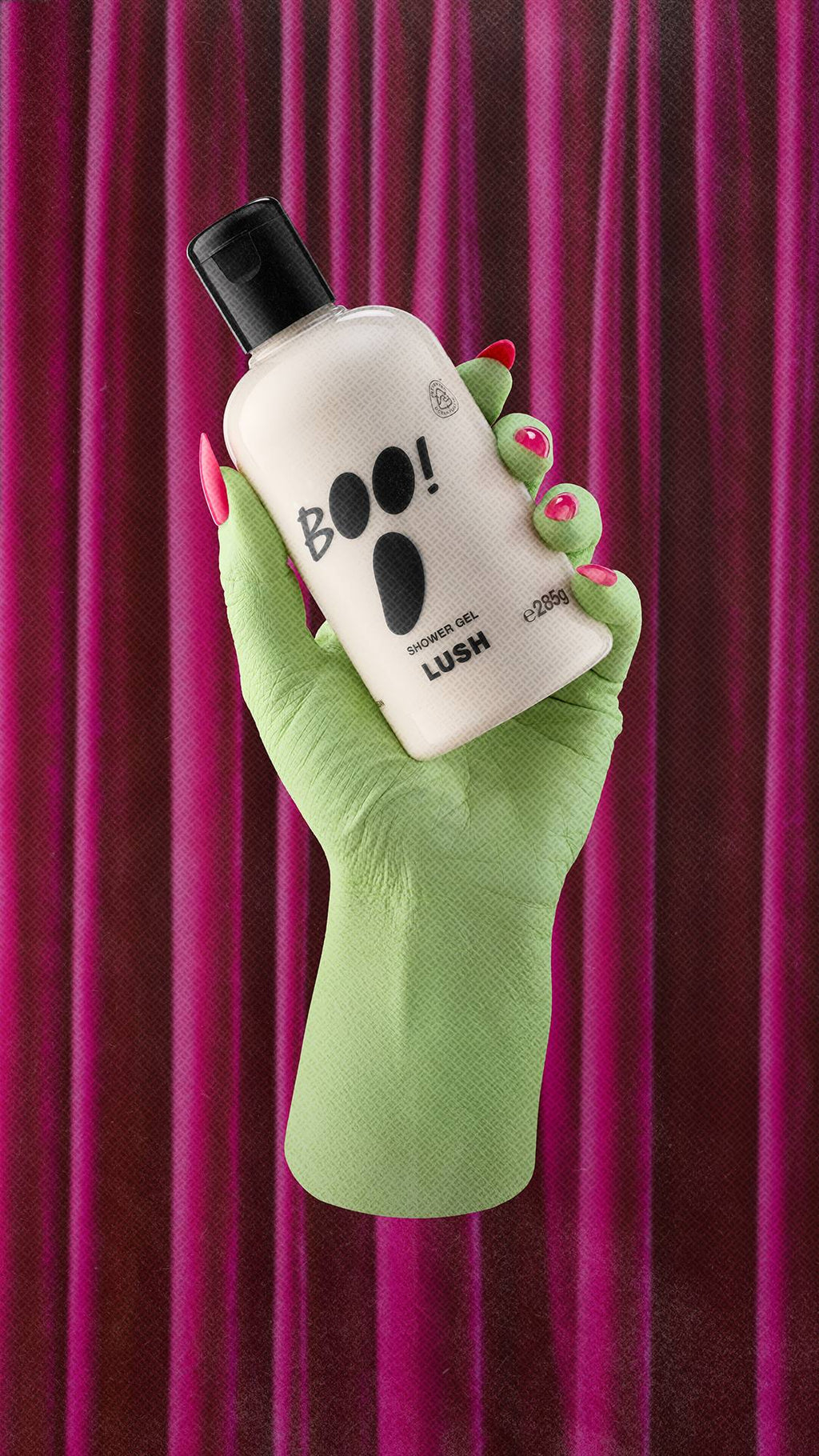 A pale green witch's hand with long, bright pink fingernails holds a bottle of Lush 'BOO!' shower gel. The bottle features a black ghost face design. The scene is set against a background of pleated, deep pink velvet curtains.