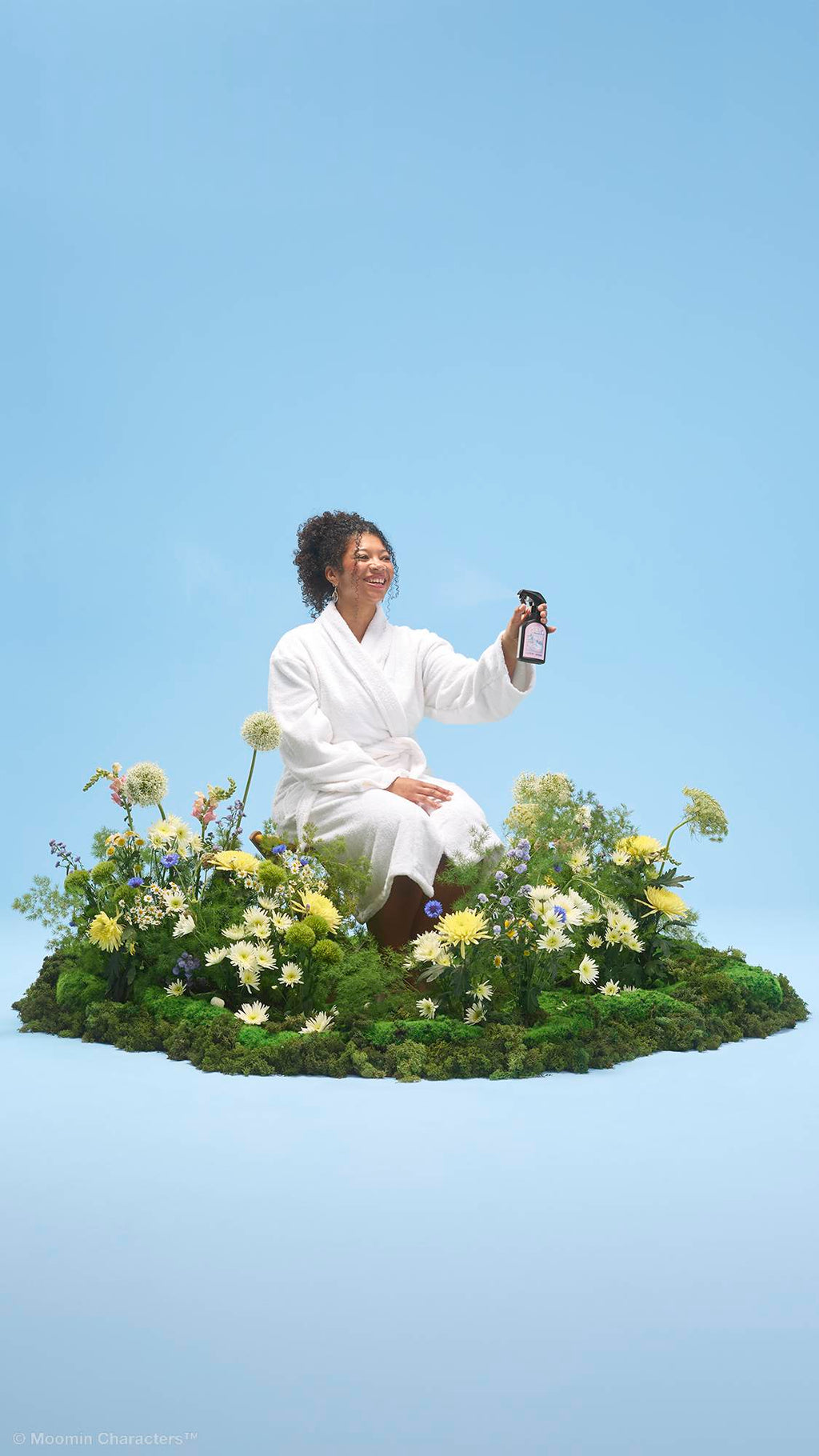 A person with curly hair wearing a white dressing gown sits on a patch of green moss and vibrant wildflowers. They are holding up a black spray bottle of "Cloudberry" body spray, all set against a solid light blue background.