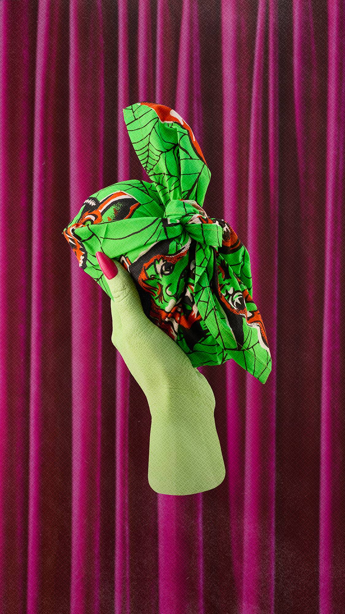 A lime green hand with a bright pink fingernail holds up a gift wrapped in a vibrant green knot-wrap fabric featuring a Halloween cobweb and character pattern. The hand is set against a background of draped magenta velvet curtains.