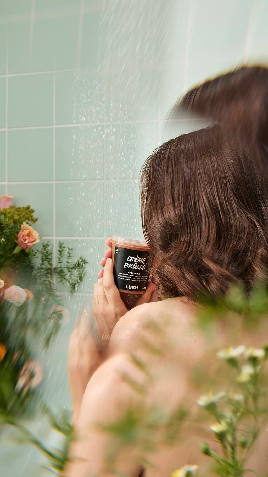 A person with wet, dark hair stands under a shower spray against a background of light green tiles, holding a black pot of "Crème Brûlée" Body Scrub. The scene is framed by blurred green foliage and flowers in the foreground.