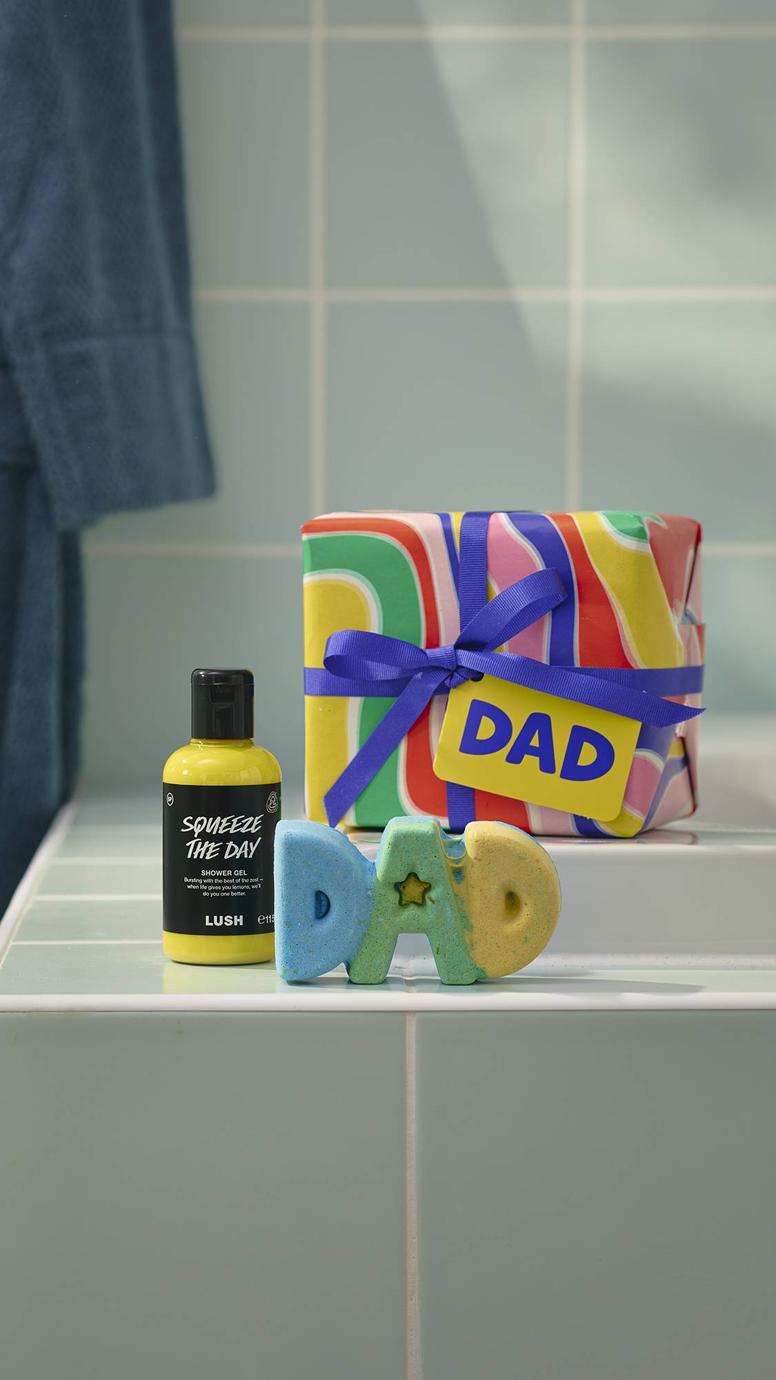 A Father's Day gift set from Lush sits on a pale green tiled ledge. It features a colourful swirl-patterned gift box with a blue ribbon, a bottle of yellow "Squeeze The Day" shower gel, and a blue and yellow bath bomb shaped like the word "DAD".
