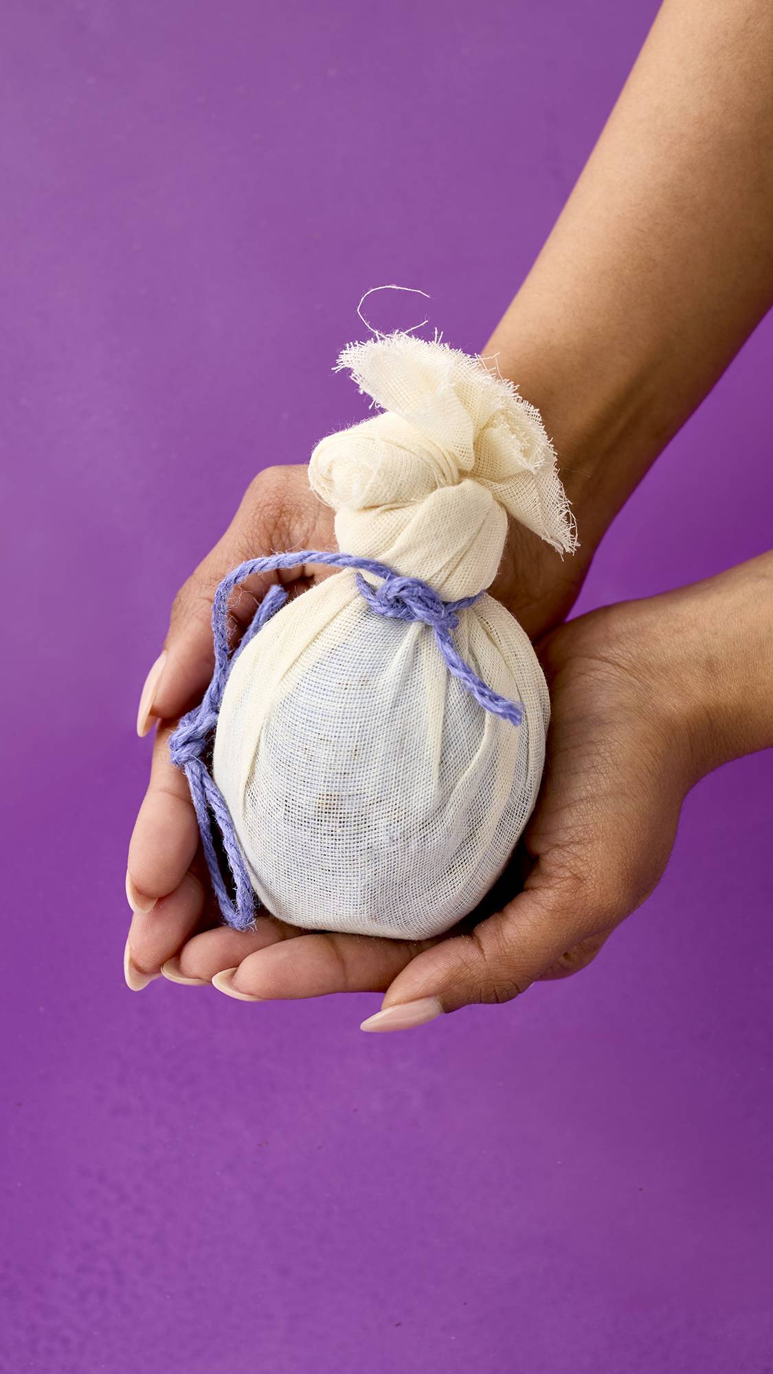 Deep Sleep. Two hands holding a spherical bath bomb wrapped in a cream-colored muslin cloth. The cloth is tied at the top with a knotted purple cord. The background is a solid, vibrant purple.
