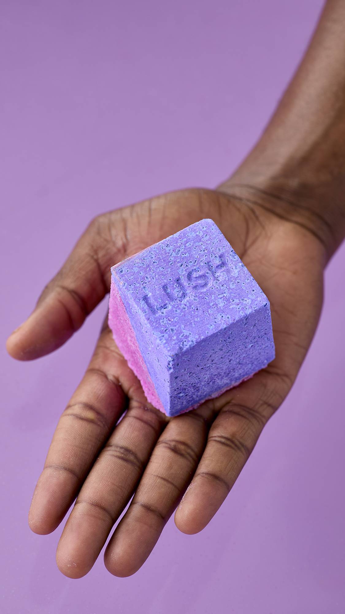 A hand holds the purple and pink Deep sleep Salt Cube bath bomb against a matching lavender background. The top surface of the cube displays a textured, embossed "LUSH" logo.