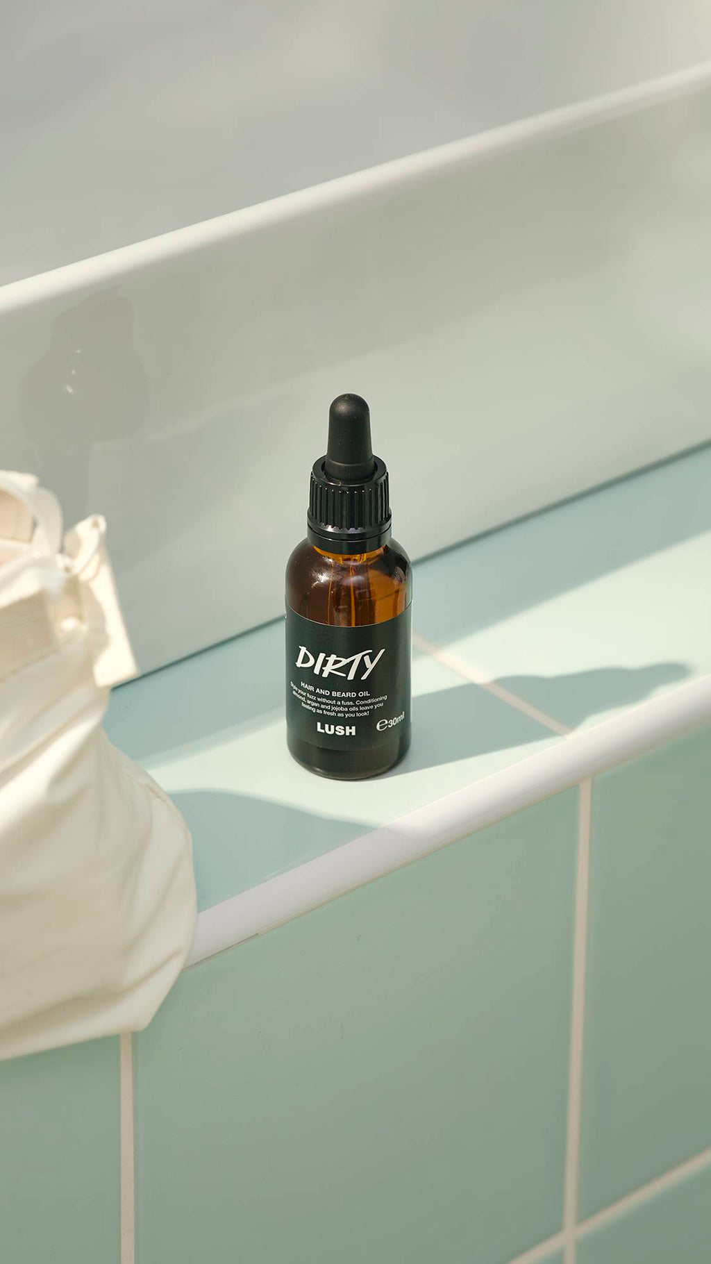 A small, amber glass bottle of "Dirty" hair and beard oil by Lush sits on a pale green tiled ledge. The bottle features a black label and a black pipette dropper lid. A white washbag is partially visible on the left.
