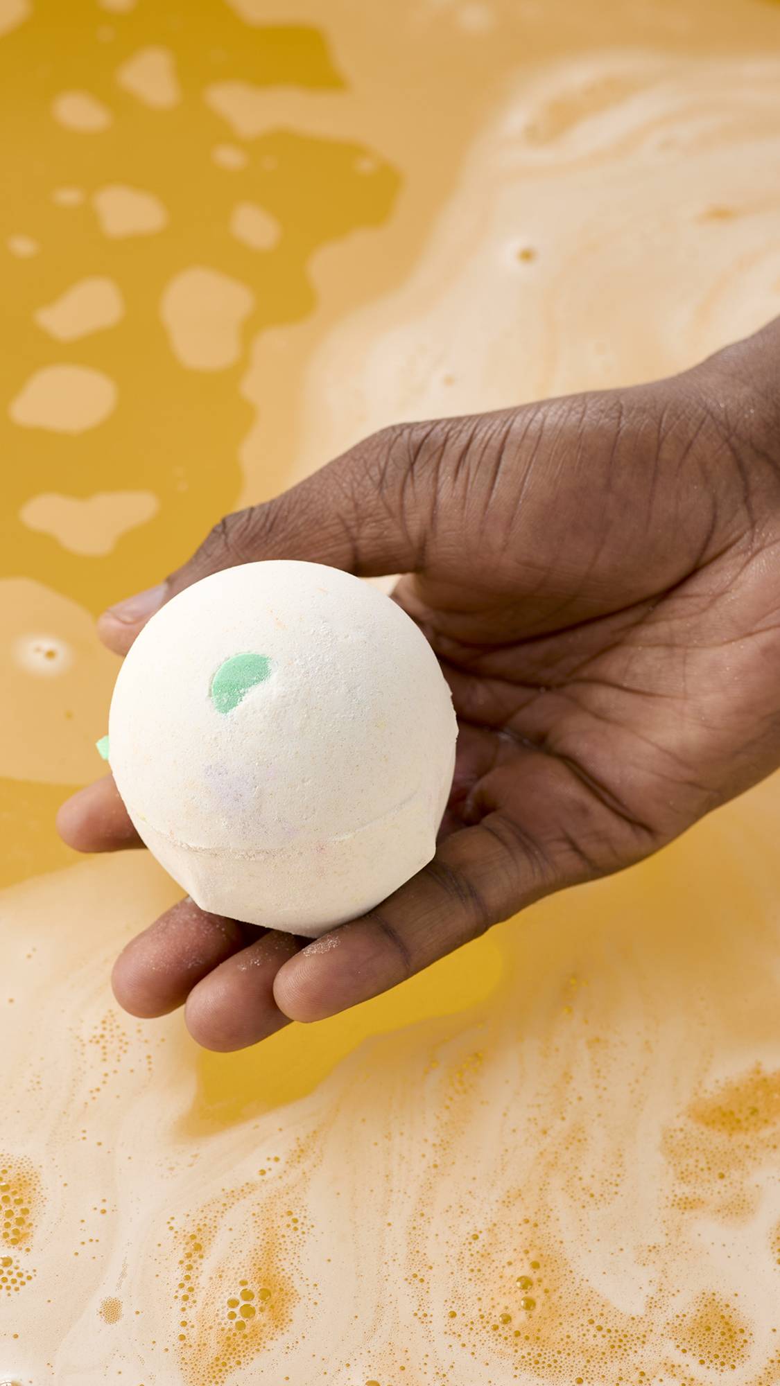 A model's hand holds a spherical, off-white "Dragon's Egg" bath bomb with small slivers of colour over a tub of swirling orange bathwater. The water is frothy with many small bubbles on the surface.