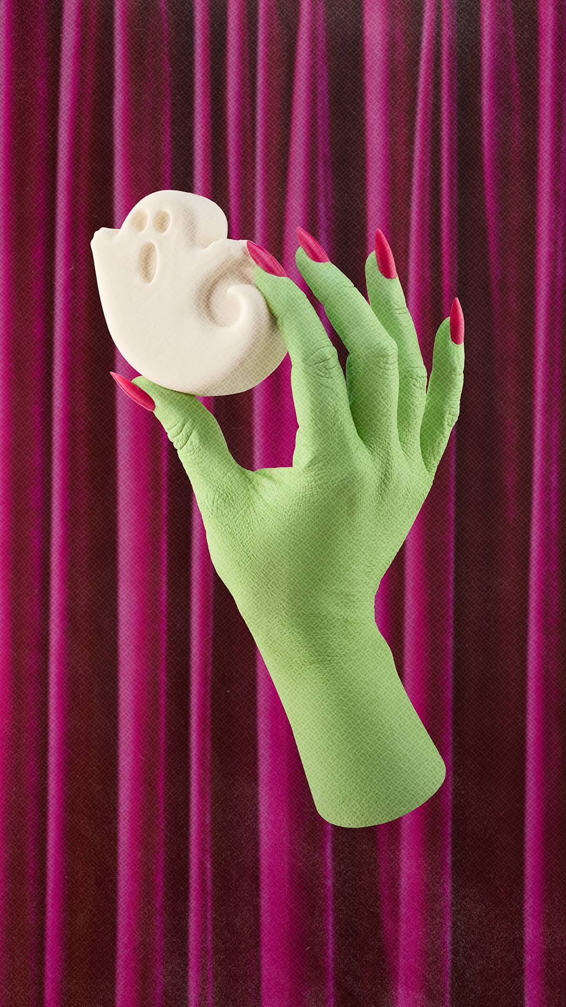 A pale green witch's hand with long, vibrant pink nails holds up an off-white bar of soap shaped like a cartoon ghost. The scene is set against a background of pleated, deep pink velvet curtains.