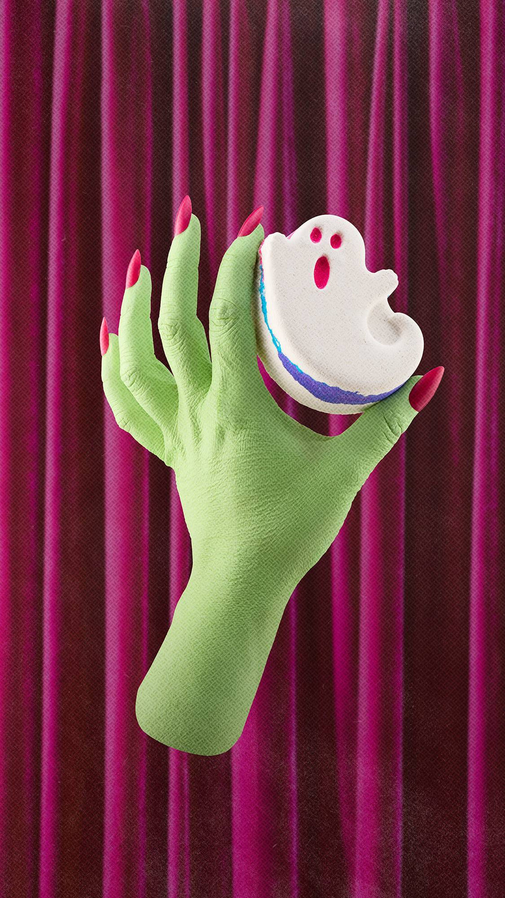 A lime green witch's hand with bright pink fingernails holds a white ghost bath product with blue and purple layered edges. The hand is set against a background of draped magenta velvet curtains.