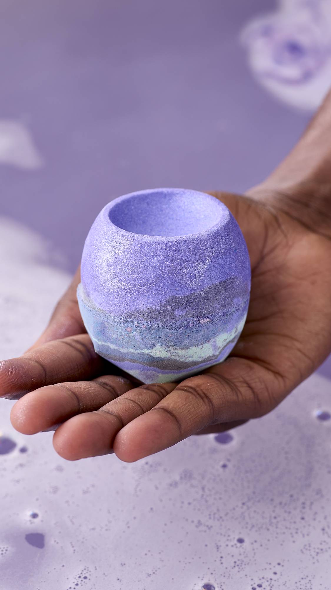 The image shows a hand holding a round, purple "Goddess" bath bomb with a grey and mint green marbled base over a tub of lilac-coloured bathwater. The bath bomb has a bowl-shaped indentation on top.