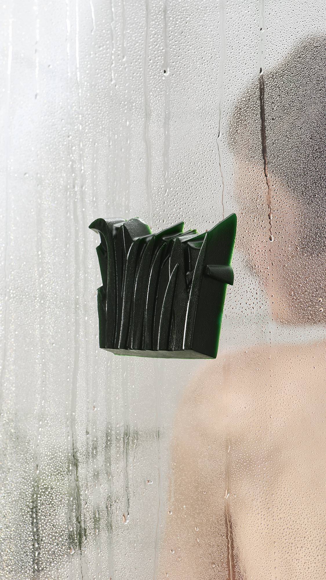 A textured, dark green shower gummy soap stuck to a steamy glass shower door covered in water droplets. A person is visible through the foggy glass in the background.