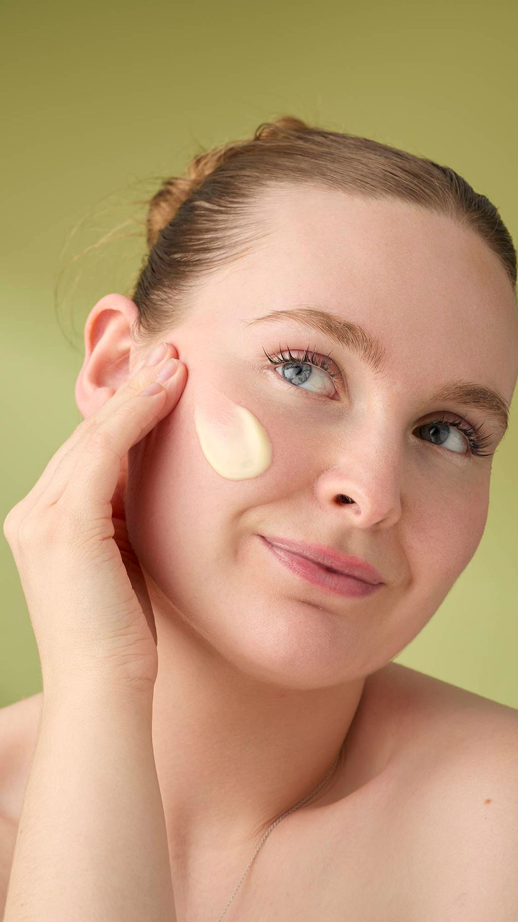 A woman with light hair smiles gently as she applies a dab of smooth Honey, Beeswax And Almond Oil facial cleanser to her cheek. She is shown from the shoulders up against a solid olive-green background that highlights the product's creamy texture.