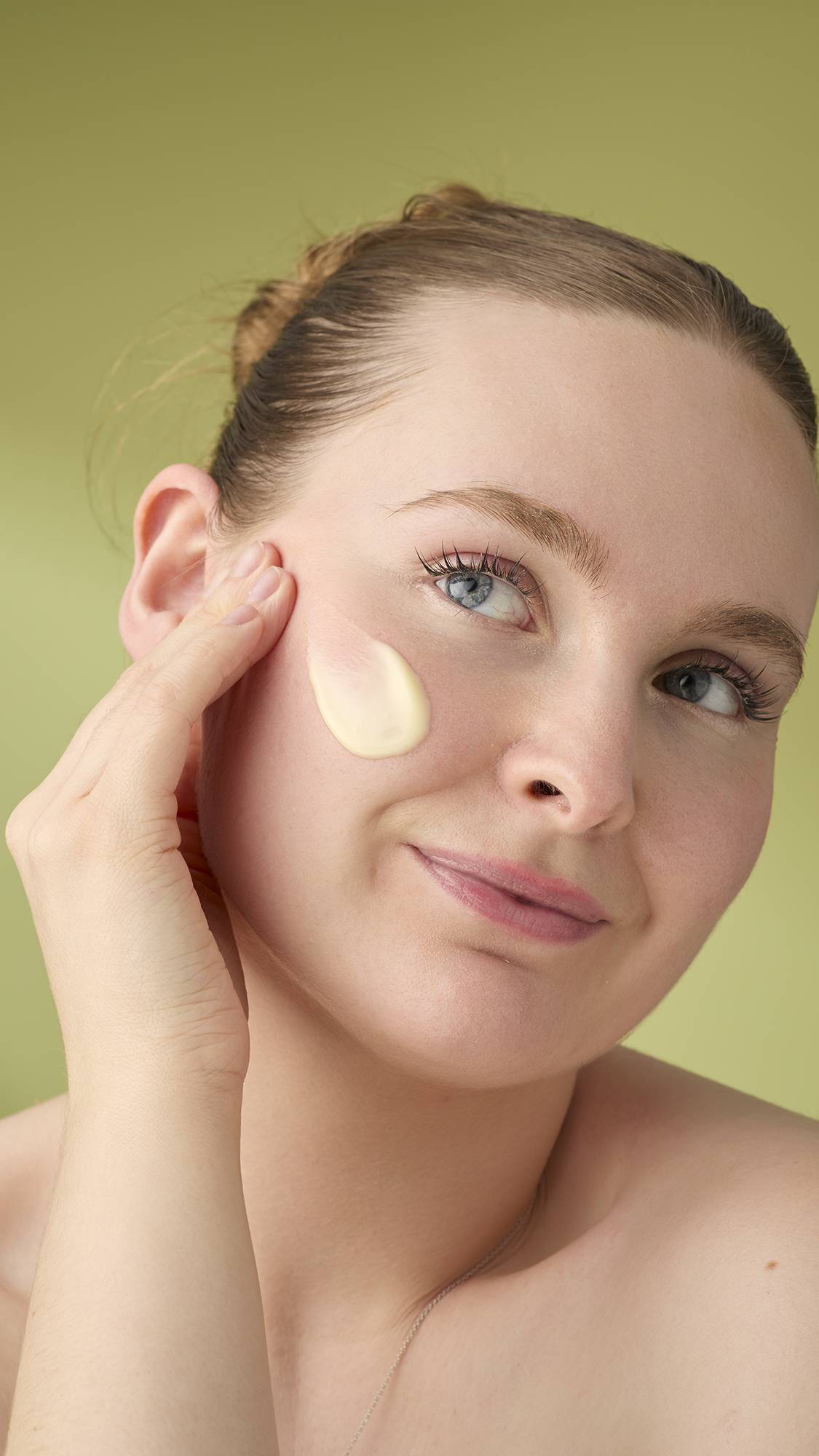 A woman with light hair smiles gently as she applies a dab of smooth Honey, Beeswax And Almond Oil facial cleanser to her cheek. She is shown from the shoulders up against a solid olive-green background that highlights the product's creamy texture.