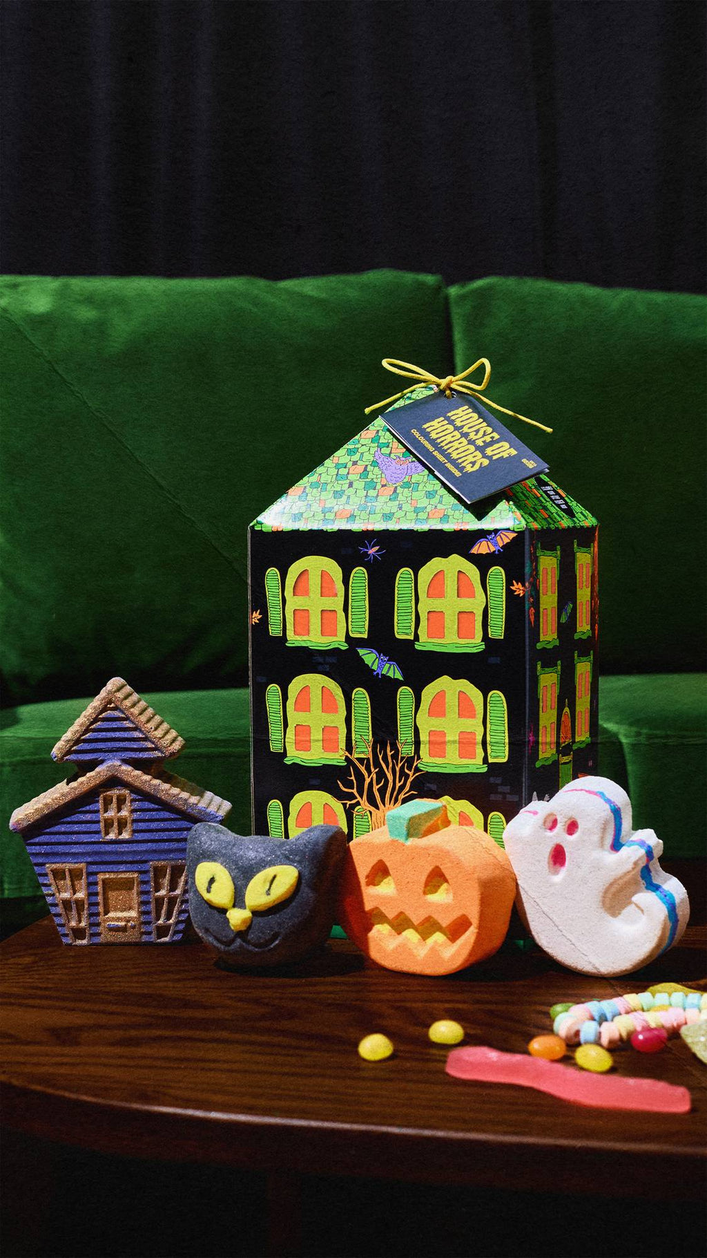 A Lush 'House of Horrors' gift box stands on a wooden table in front of a green velvet couch. Arranged in front of it are a purple house, a black cat, an orange pumpkin, and a white ghost bath product, alongside colourful sweets.