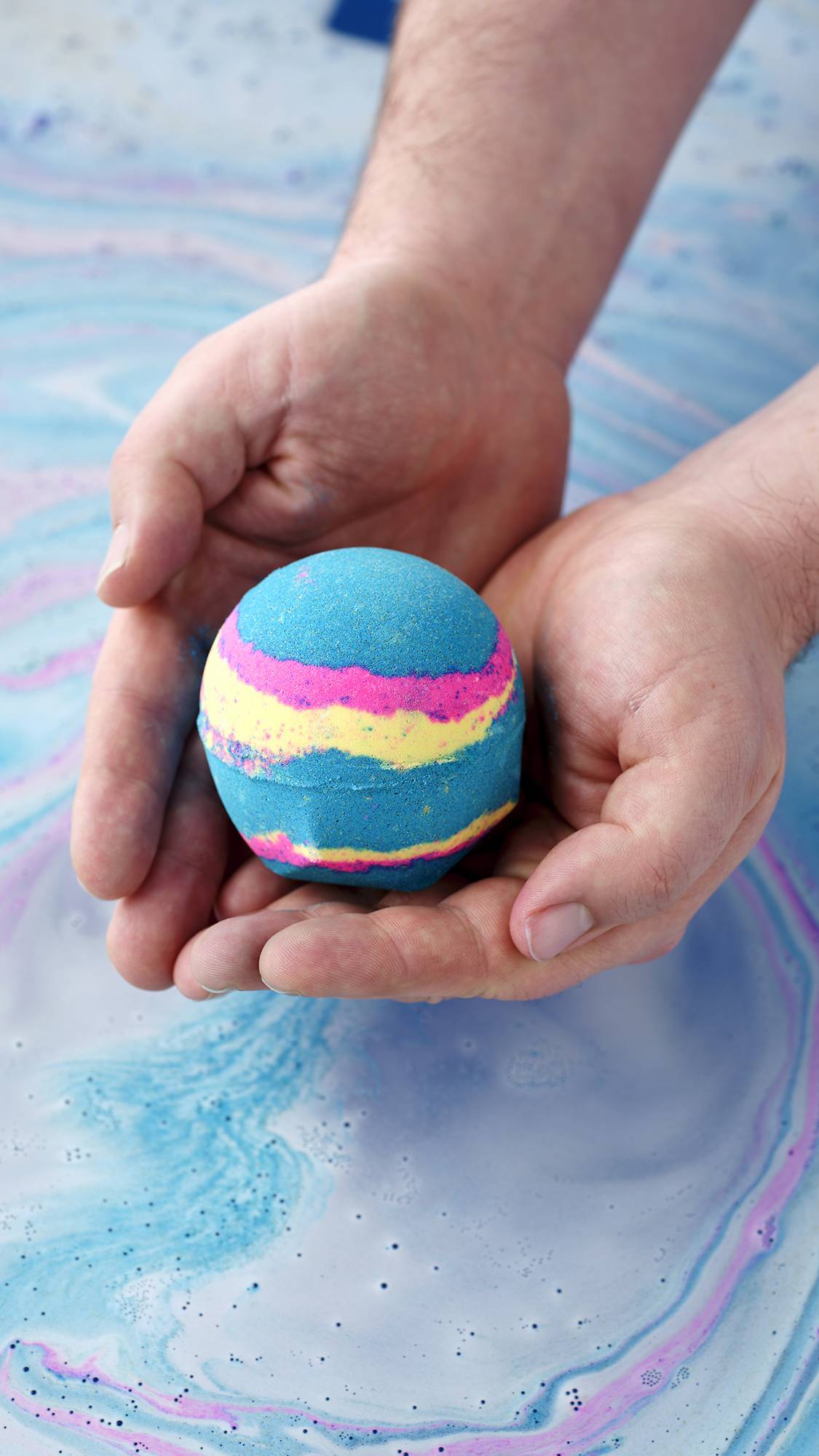 A pair of hands holds a spherical Intergalactic bath bomb over blue and pink swirling water. The bomb is primarily bright blue with vibrant horizontal stripes of neon pink and yellow.