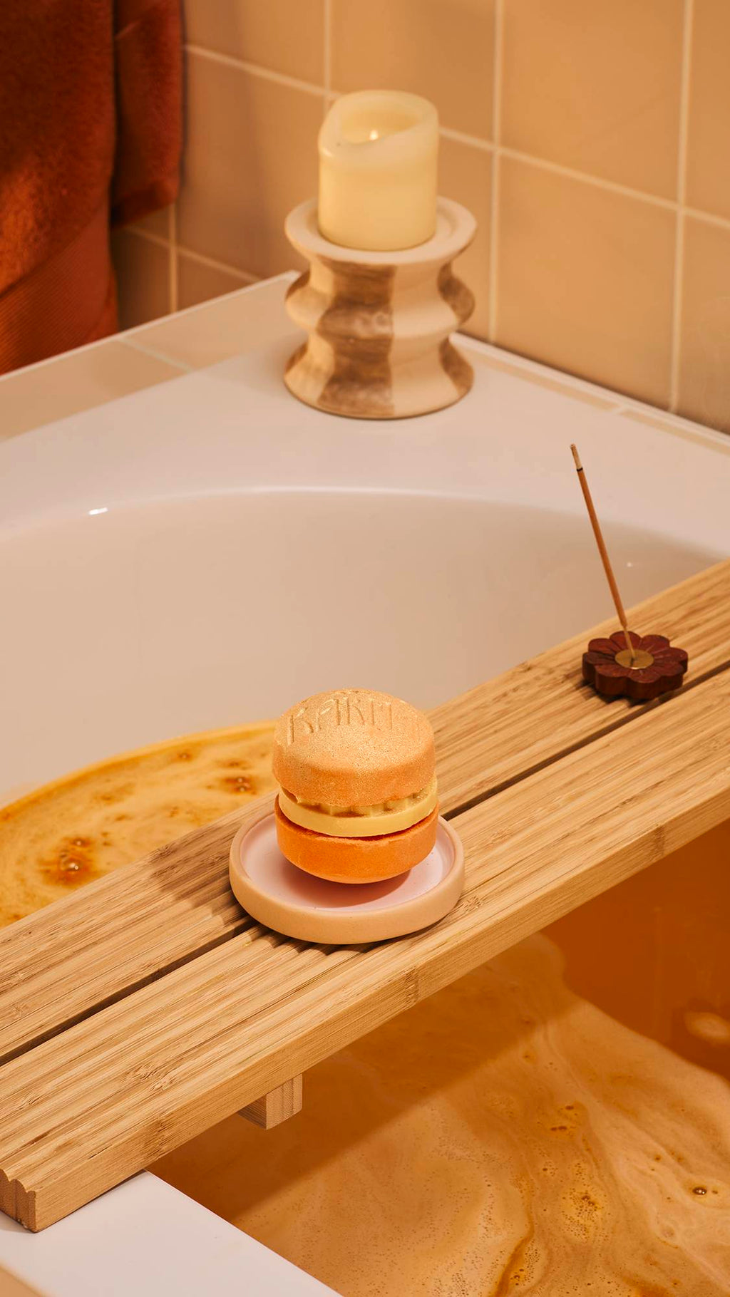 A shimmering orange 'Karma' macaron-style bath bomb sits on a small ceramic dish atop a wooden bath tray. In the background, a lit candle and a stick of incense provide a relaxing atmosphere against warm, beige bathroom tiles.