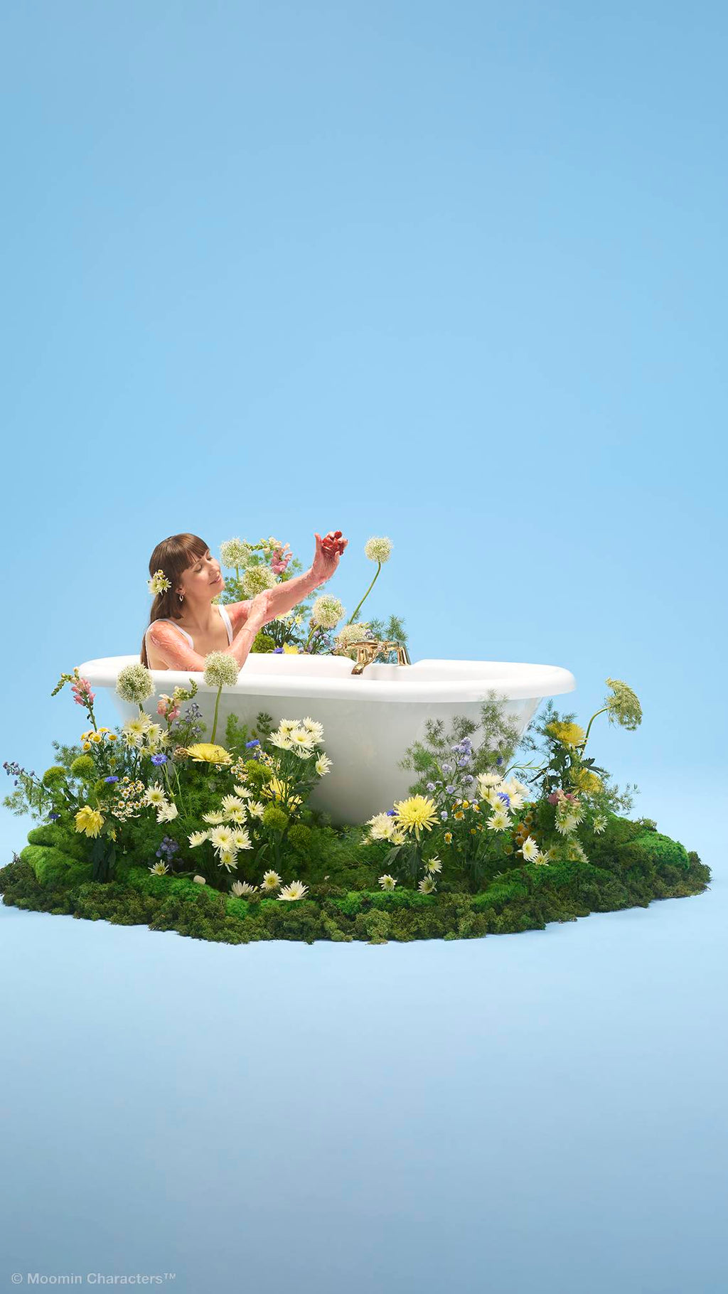 A person sits inside a white bathtub, washing their arm with a Lush "Little My" character-shaped soap. The tub is placed on a patch of lush green moss overflowing with yellow and white wildflowers, set against a plain light blue background.