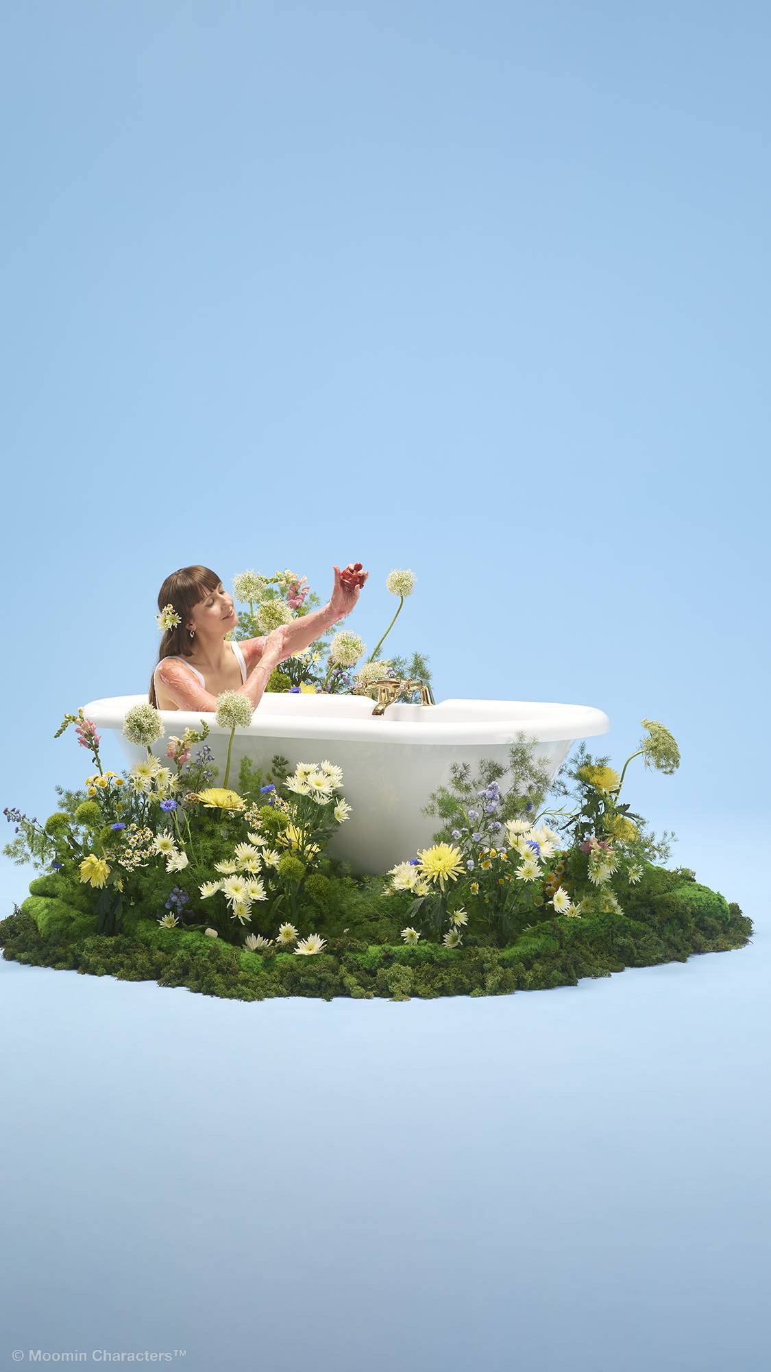 A person sits inside a white bathtub, washing their arm with a Lush "Little My" character-shaped soap. The tub is placed on a patch of lush green moss overflowing with yellow and white wildflowers, set against a plain light blue background.