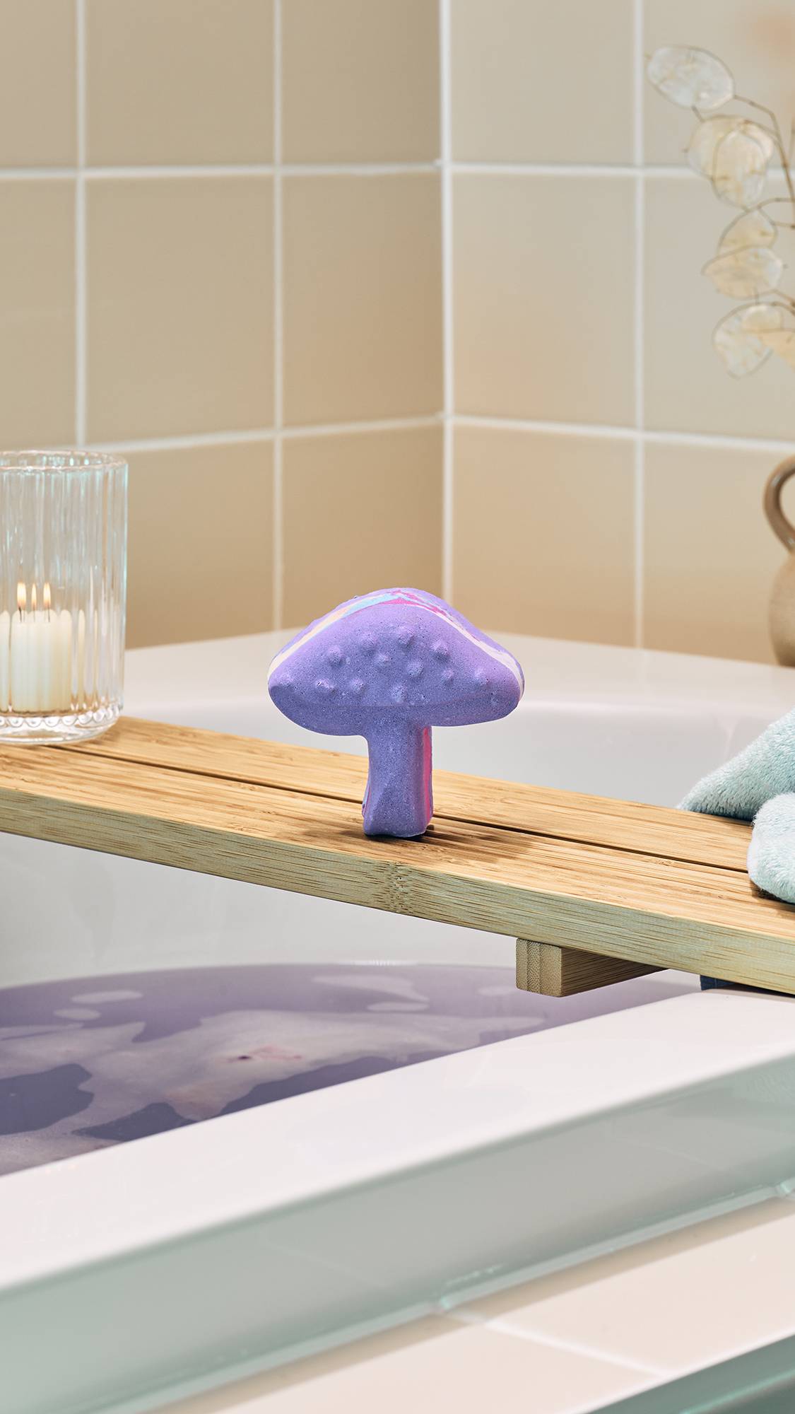 A purple "Magik Mushroom" bath bomb sits upright on a wooden bath tray over a tub of lilac-coloured water. In the background, beige tiles and a lit candle in a glass holder create a serene atmosphere.