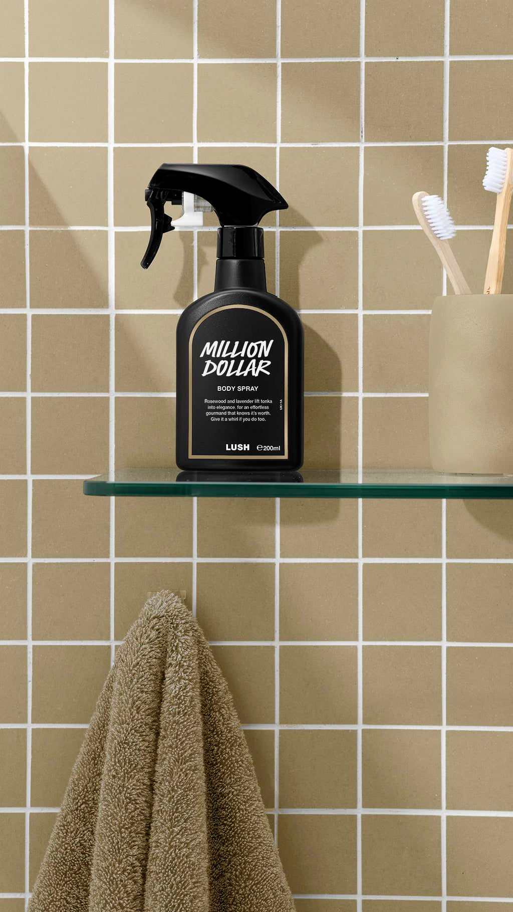A black bottle of Lush 'Million Dollar' body spray sits on a glass shelf against a beige tiled wall. To the right is a pale cup holding toothbrushes, and below the shelf hangs a fluffy, beige towel.