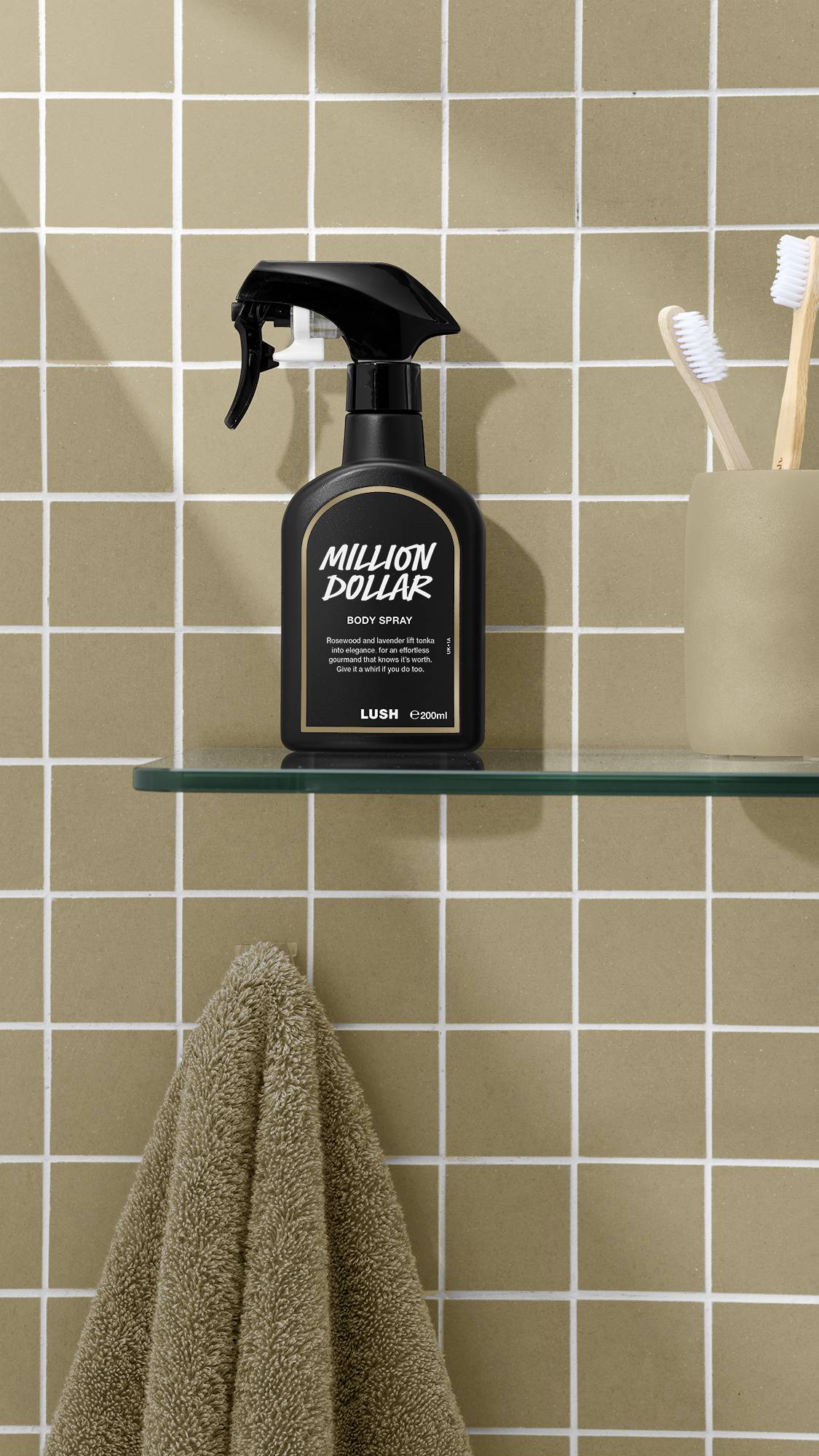 A black bottle of Lush 'Million Dollar' body spray sits on a glass shelf against a beige tiled wall. To the right is a pale cup holding toothbrushes, and below the shelf hangs a fluffy, beige towel.