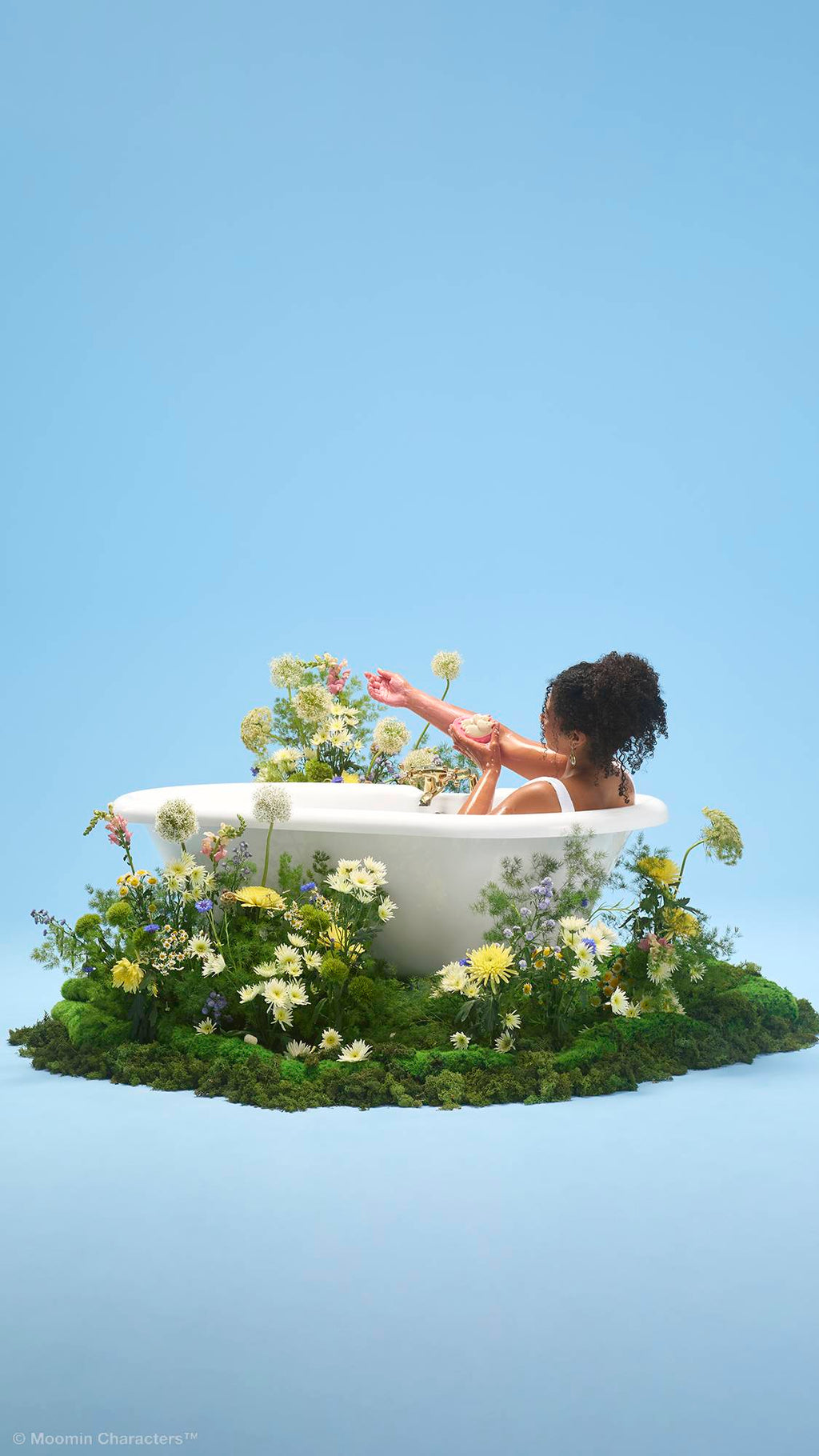 A person with curly hair sits in a white bathtub, washing their arm with a pink and white "Moomin Delight" soap and body wash. The tub sits on a patch of green moss filled with wildflowers, set against a plain light blue background.