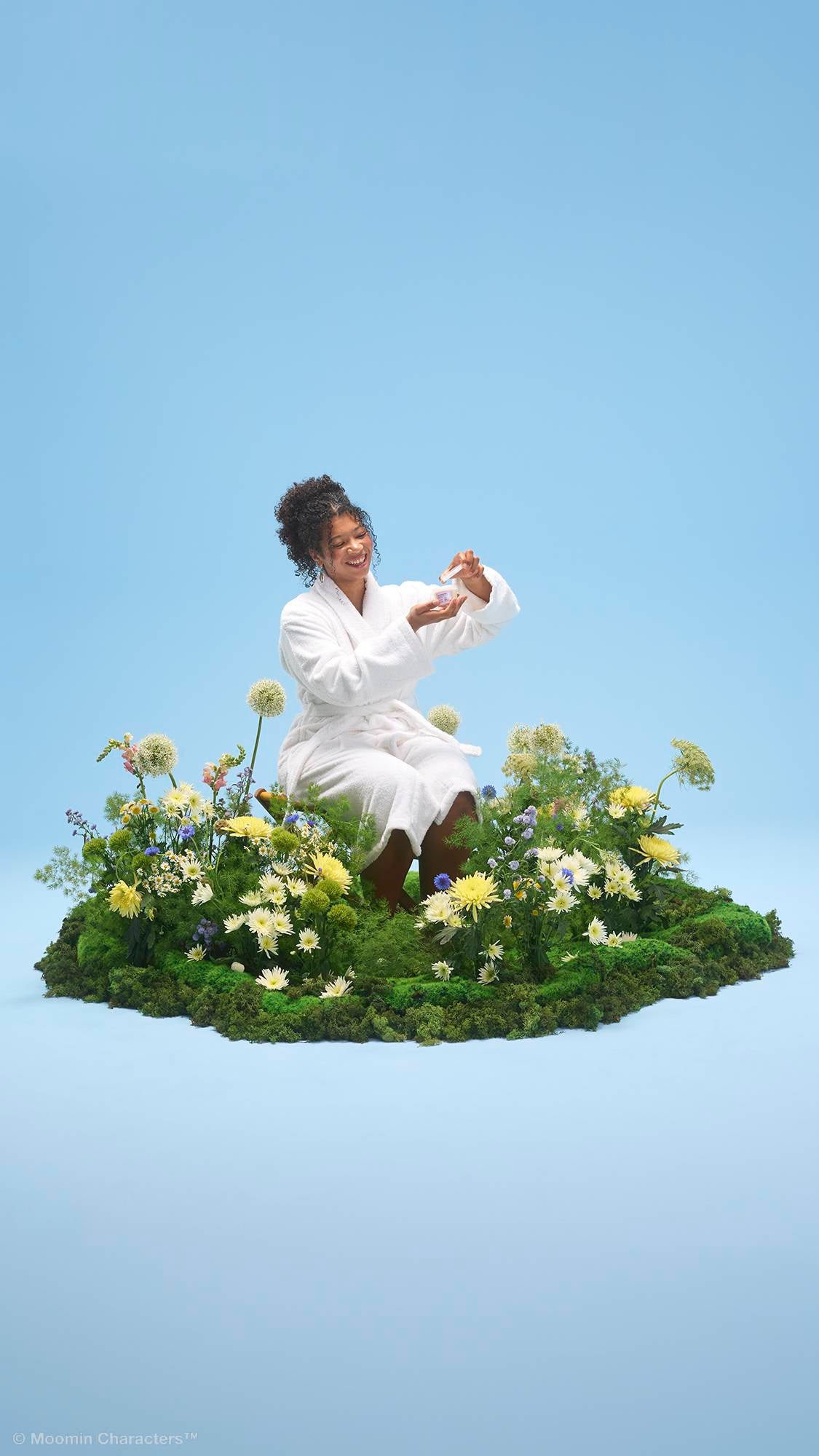 A smiling person wearing a white dressing gown sits amongst a lush patch of green moss and vibrant yellow and white wildflowers. They are holding open a small tub of Moominmamma hand and body lotion, all set against a solid light blue background.