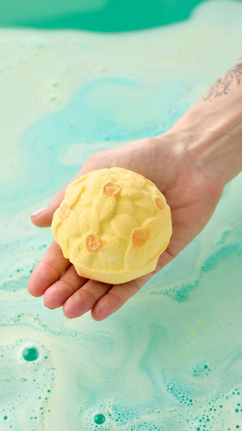 A hand holds a pale yellow, floral-patterned bath bomb with shimmering gold centers over a bath of light green water. Swirls of pale blue and yellow foam create a marbled texture across the water's surface.