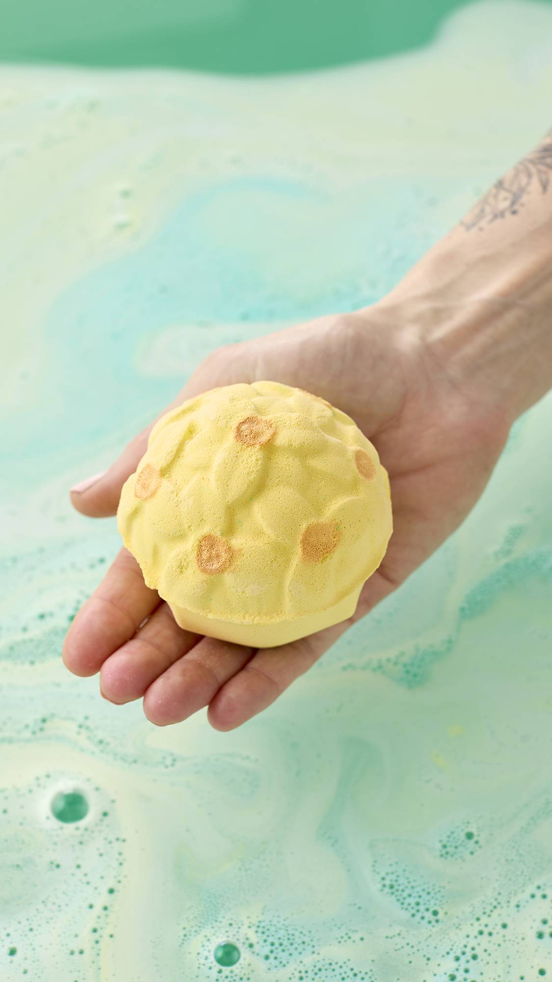 A hand holds a pale yellow, floral-patterned bath bomb with shimmering gold centers over a bath of light green water. Swirls of pale blue and yellow foam create a marbled texture across the water's surface.