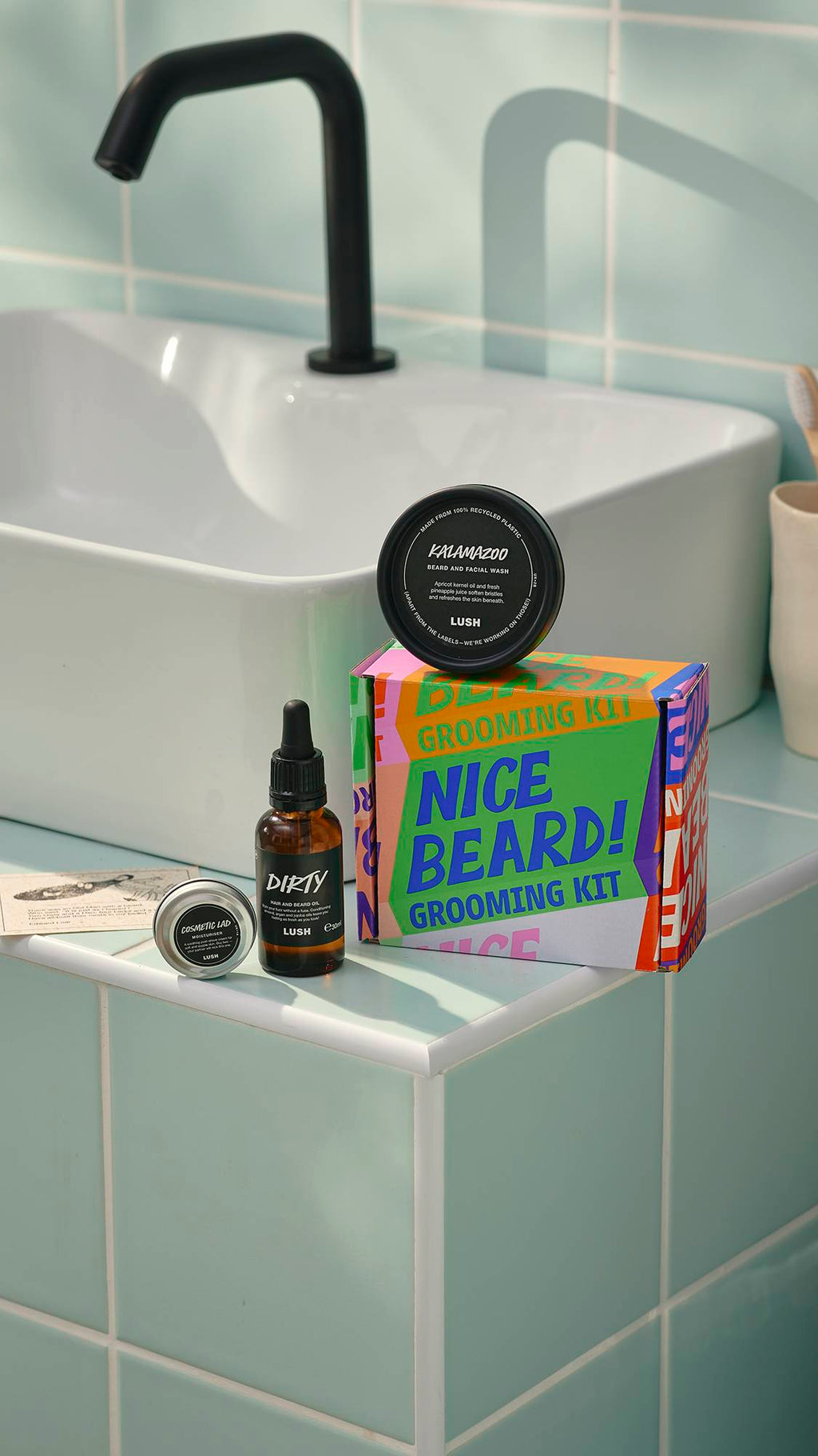 A Lush ‘Nice Beard!’ grooming kit sits on a tiled bathroom ledge next to a white sink. The set includes a colourful typographic box, a bottle of Dirty hair and beard oil, a tub of Kalamazoo beard wash, and a small tin of Cosmetic Lad moisturiser.