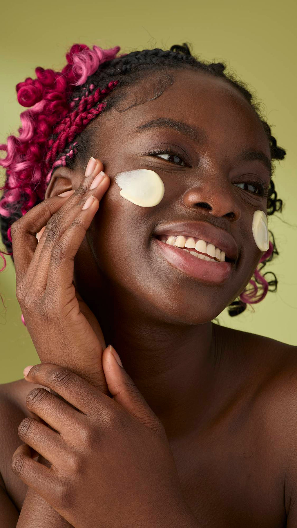 A woman with pink and black braids smiles as she applies a smooth, ivory Oat facial cleanser to her cheeks. The thick, creamy product stands out against her skin and the solid olive-green background, emphasising its rich texture.