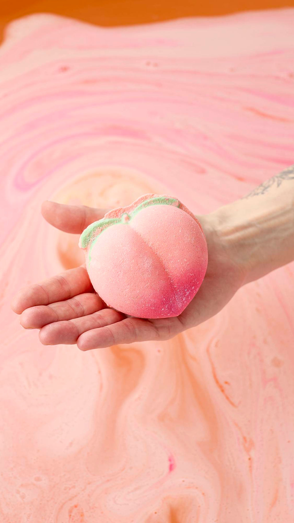 A hand holds a peach-shaped bath bomb over a tub of swirling peach and pink bathwater. The bath bomb has a soft pink gradient and small green leaves at the top. The water surface is covered in delicate foam and vibrant streaks of colour.