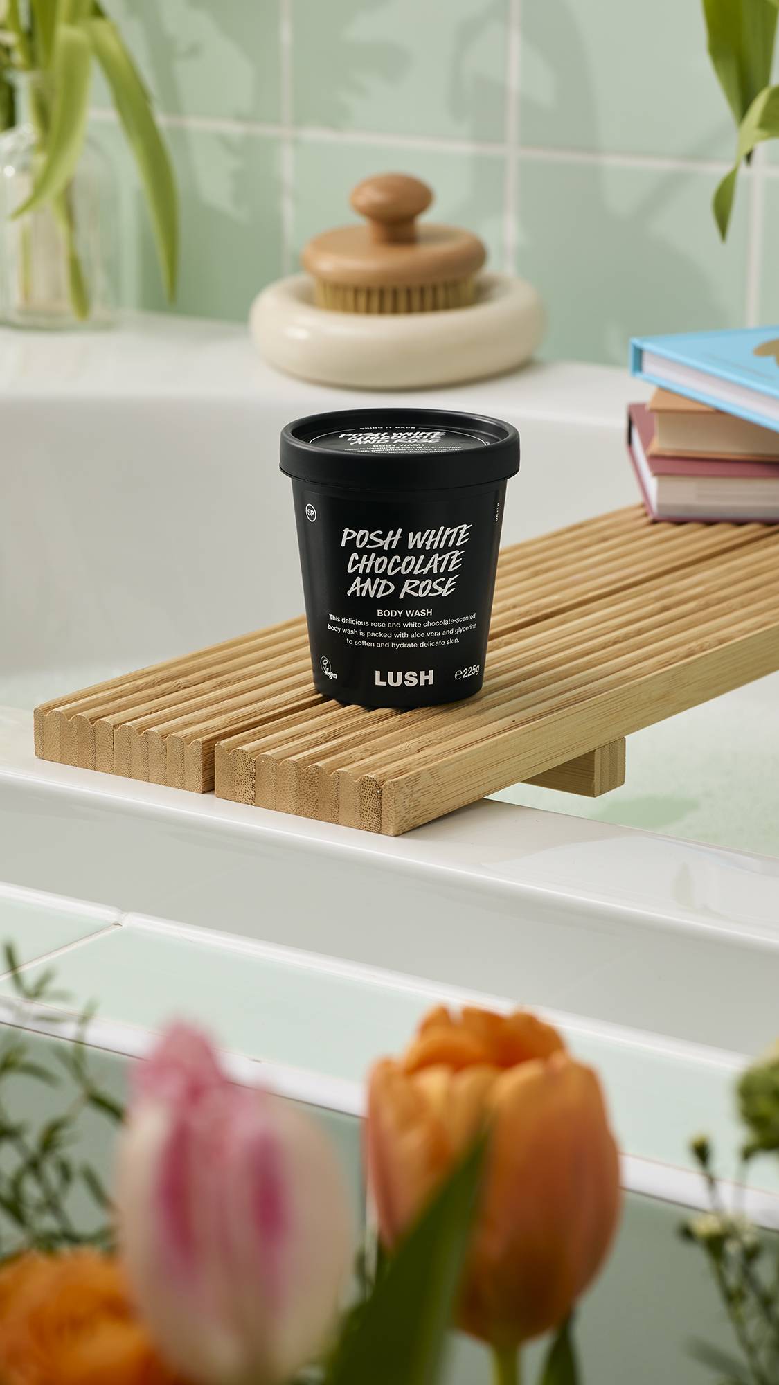 A black pot of "Posh White Chocolate and Rose" Body Wash sits on a wooden bath tray. The label notes it contains aloe vera and glycerine. The scene is set against mint green tiles with blurred tulips in the foreground.