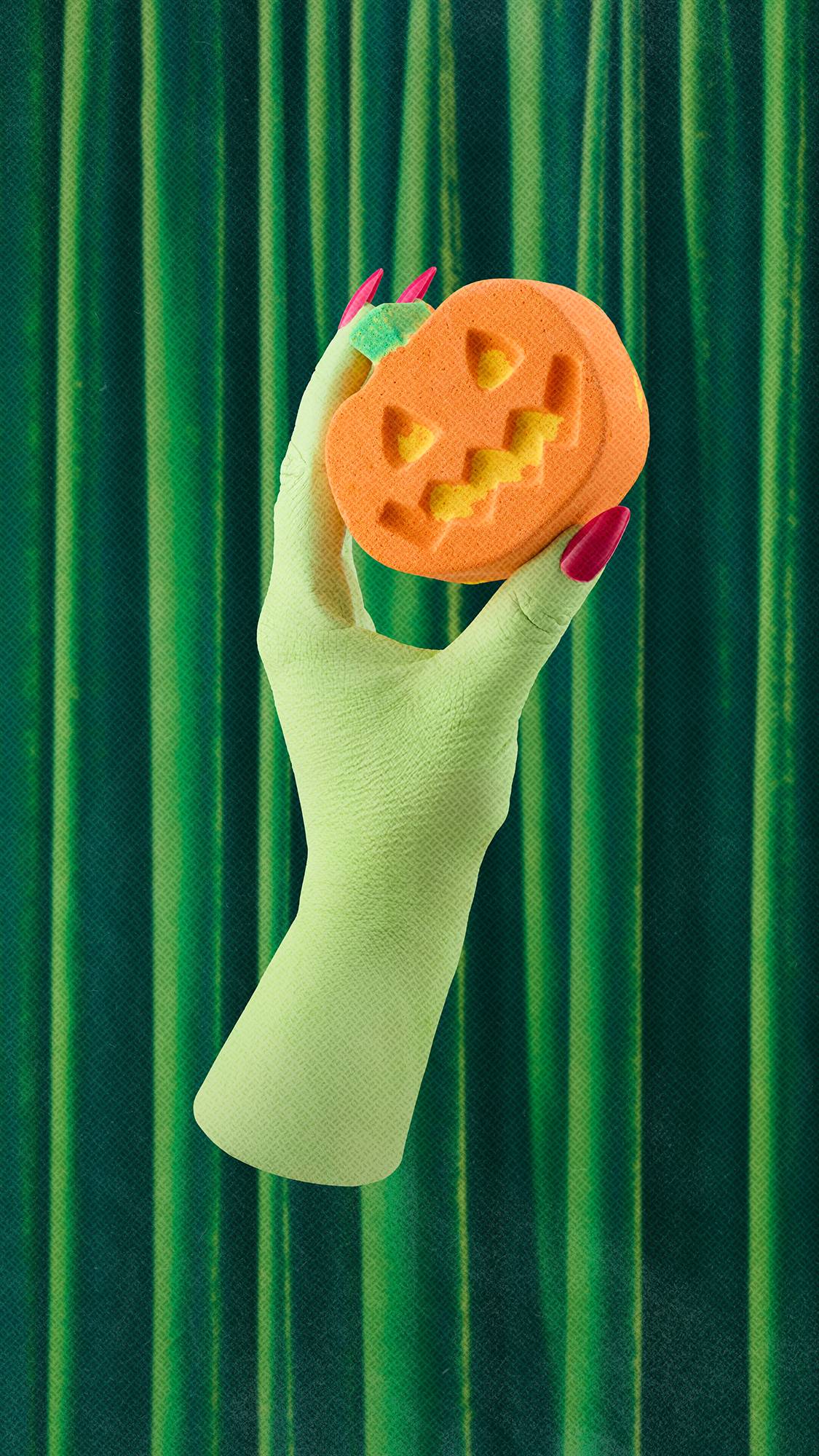 A hand holds an orange pumpkin-shaped bath bomb over a bath. The pumpkin features carved-out eyes and a jagged smile revealing a yellow interior, and a small green stalk. The bath water below is a swirling, opaque pastel orange.