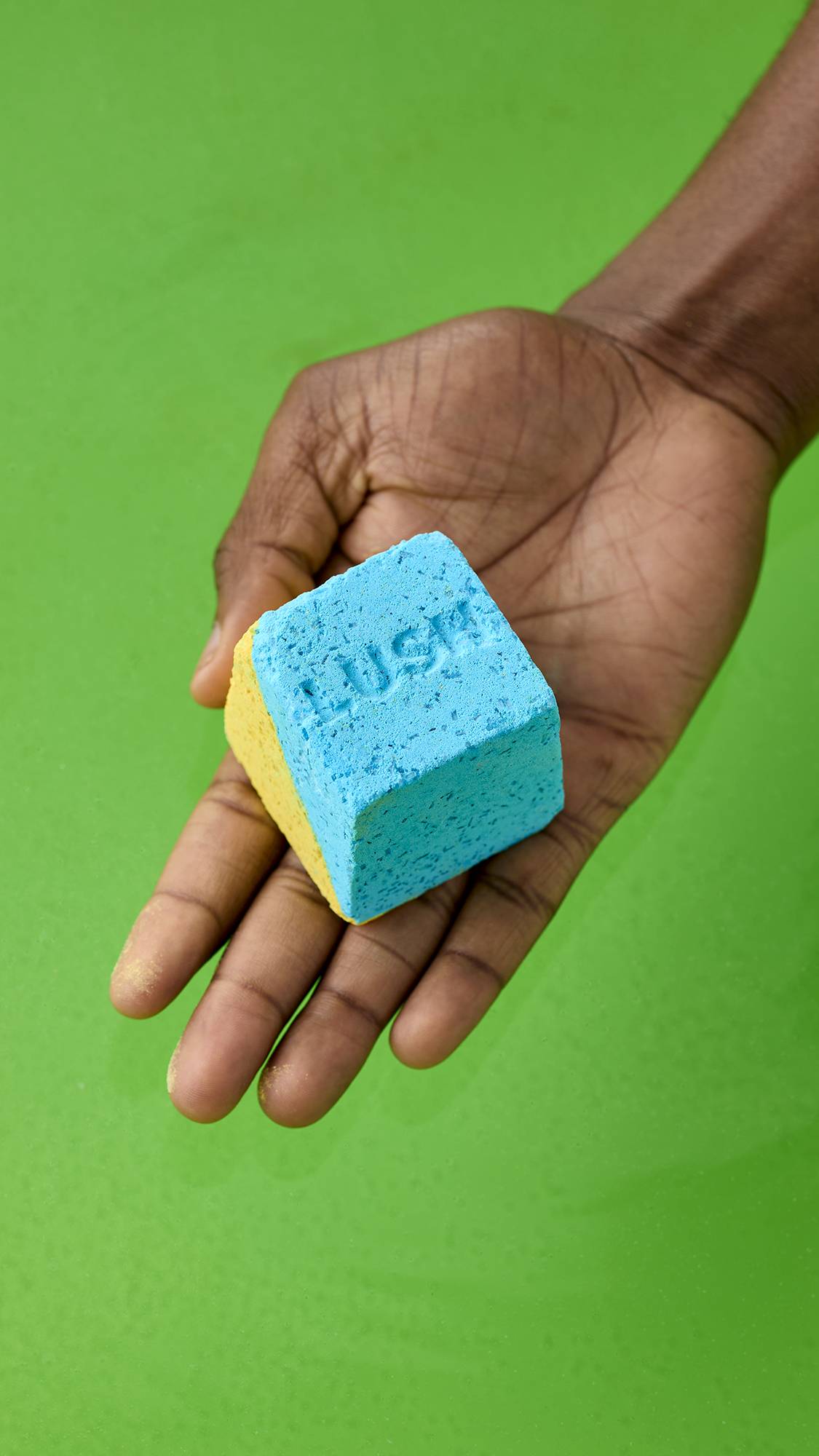 An open palm holds a cube-shaped, dual-coloured "Salt Water Soother" bath bomb in bright blue and yellow. The blue face has the word "LUSH" debossed on it, above deep green bathwater.
