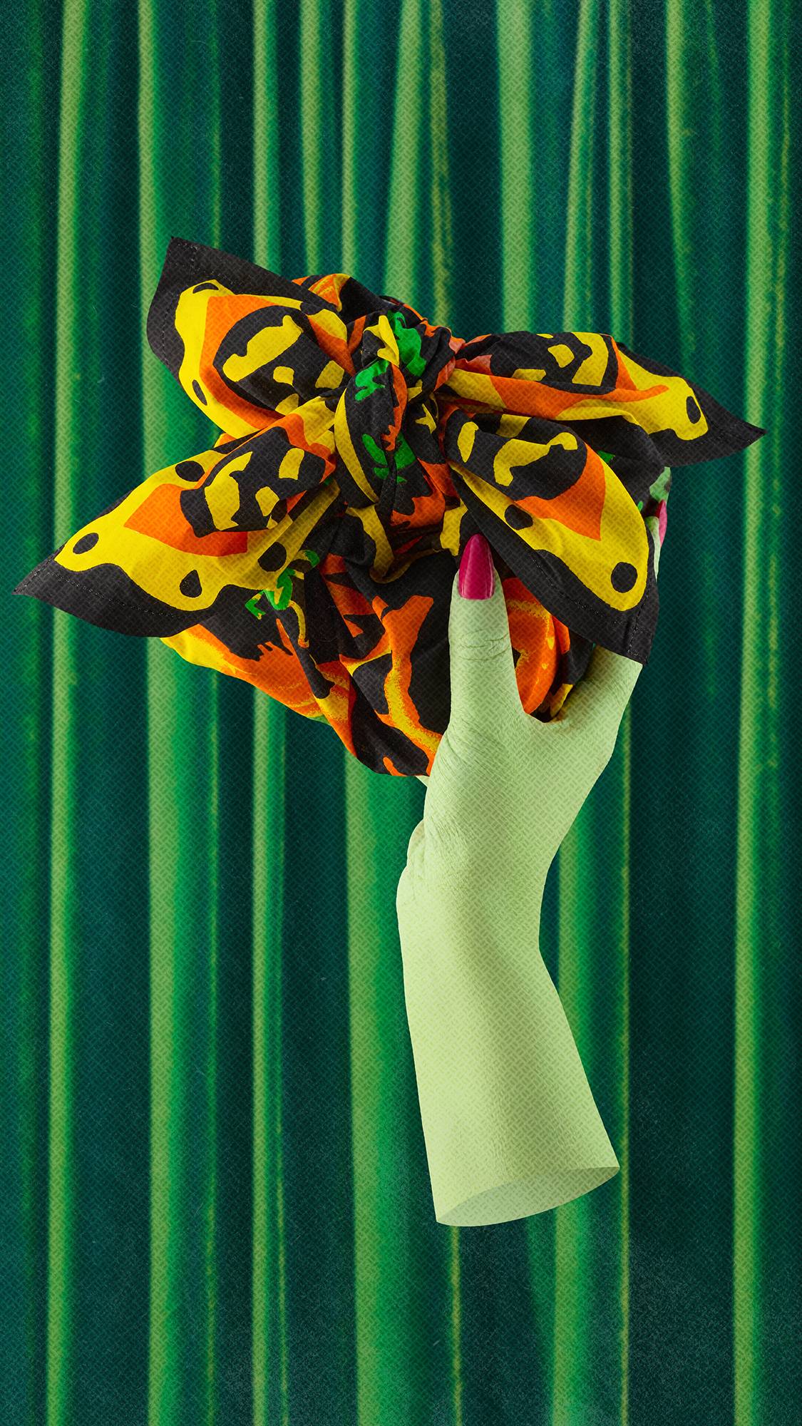A lime green hand with a bright pink fingernail holds up a gift wrapped in a colourful fabric fashioned from the "Season of the Witch" knot wrap. The hand is set against a background of draped green velvet curtains.