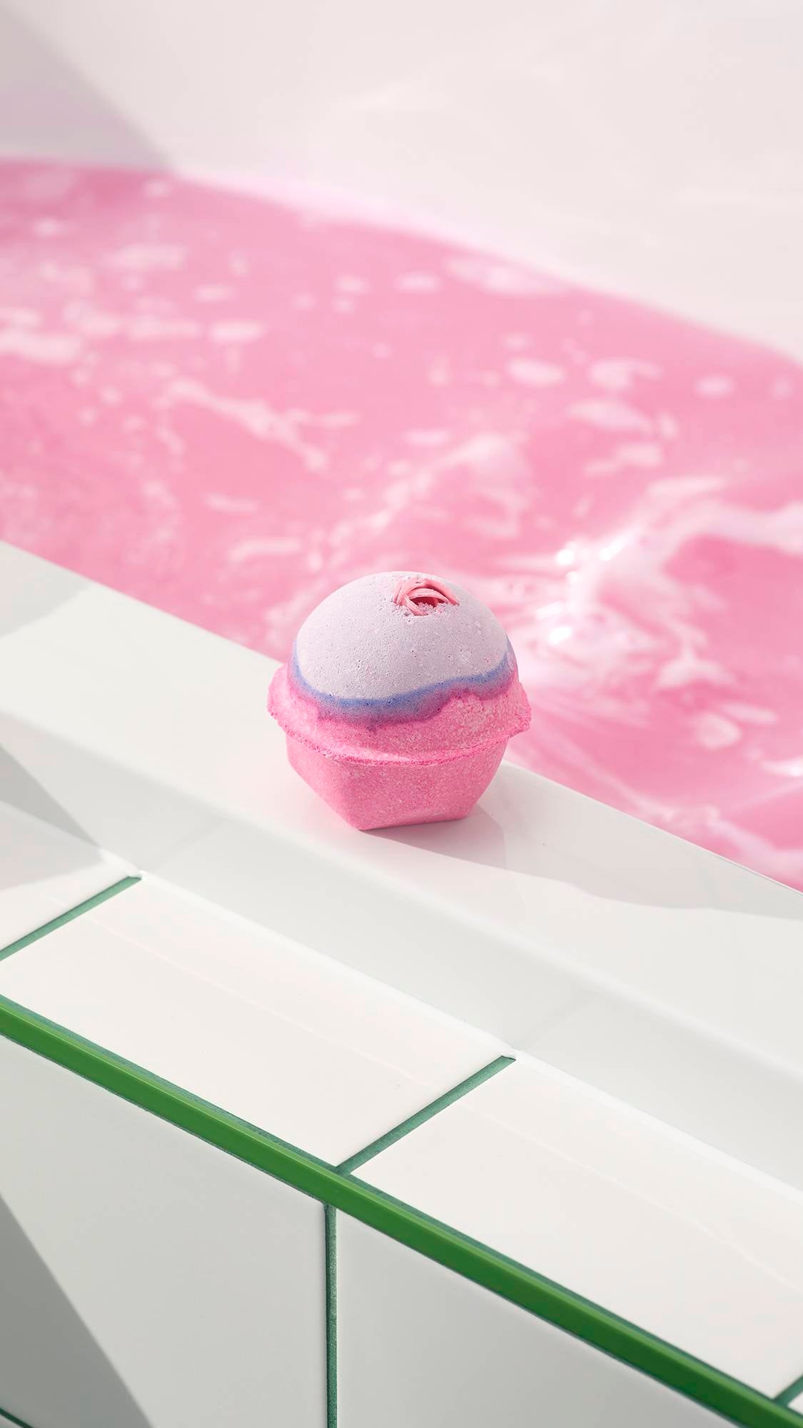 The Sex Bomb Bath Bomb, featuring a pink and purple design with a small rosebud, sits on the edge of a bathtub filled with frothy pink water.
