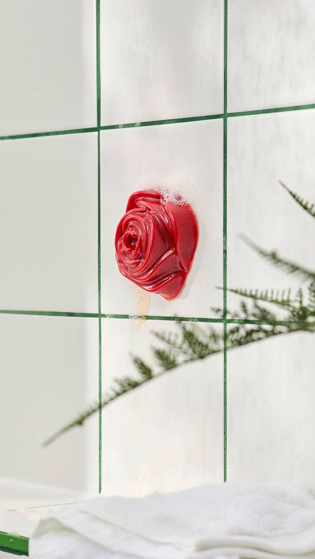 A red, rose-shaped shower gummy soap stuck to white bathroom tiles with green grout lines and soapy foam. Out-of-focus fern leaves appear in the foreground.