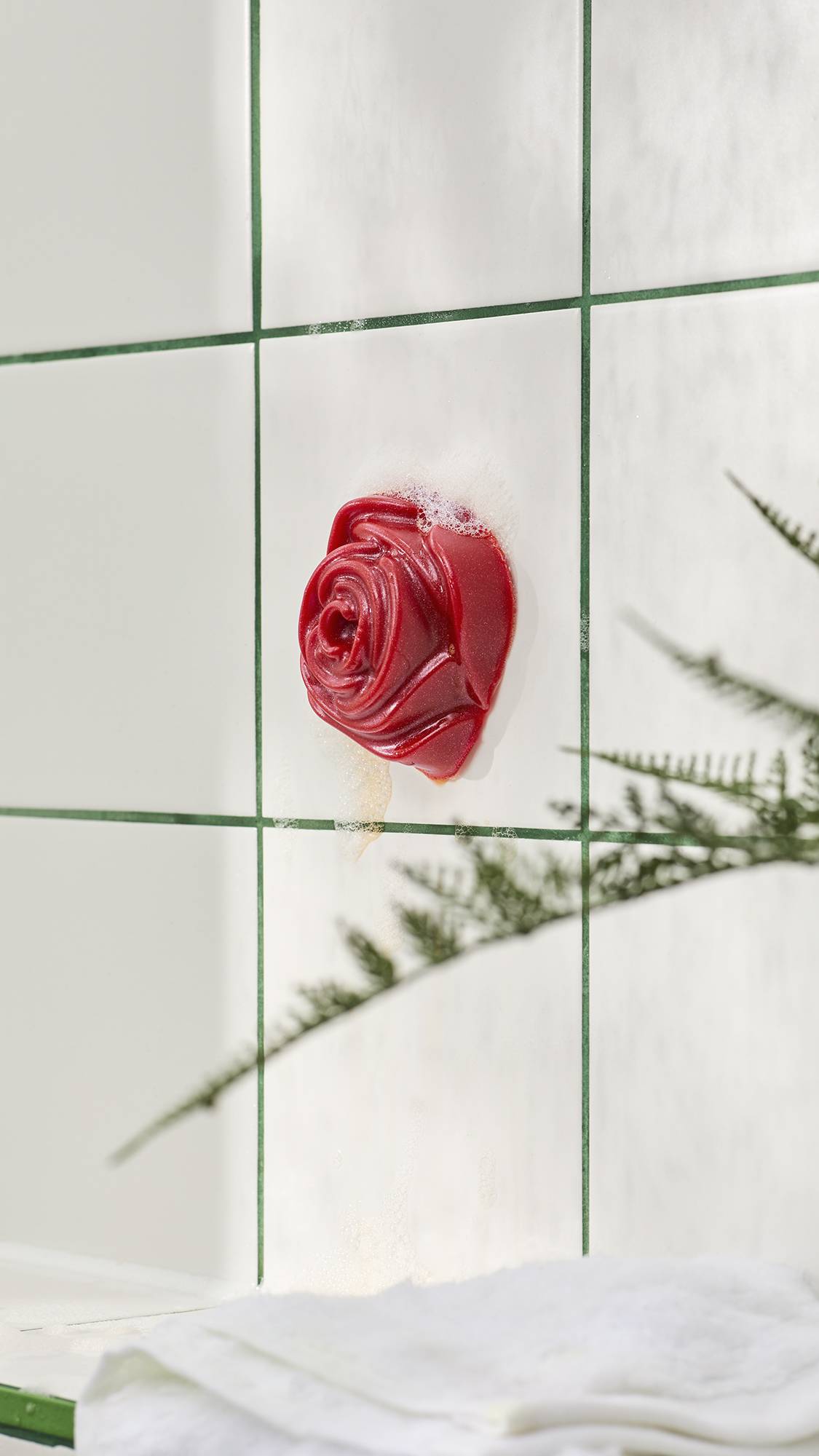A red, rose-shaped shower gummy soap stuck to white bathroom tiles with green grout lines and soapy foam. Out-of-focus fern leaves appear in the foreground.