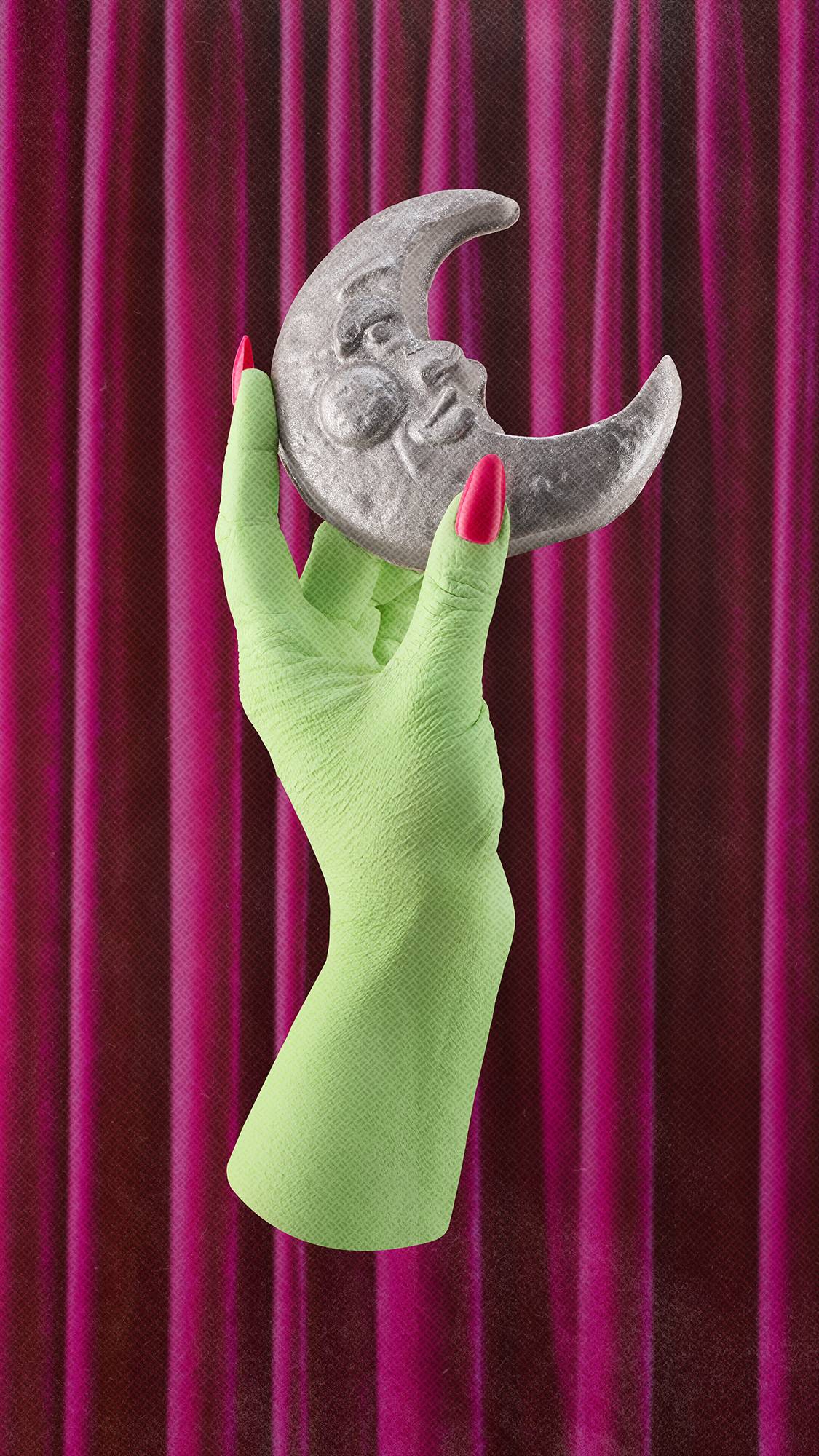 A lime green witch's hand with bright pink fingernails holds a silver, textured crescent moon soap with a stylised face, set against a background of draped magenta velvet curtains.