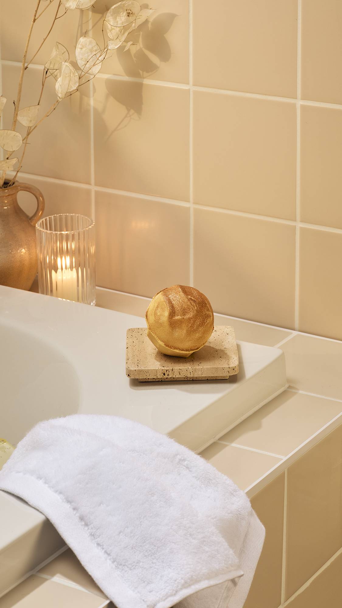 A gold, "Sticky Dates" bath bomb with a shimmering, metallic finish sits on a stone dish. In a warm-toned bathroom, it is positioned on the edge of a white bathtub near a lit candle, a white flannel, and dried flowers in a rustic vase.