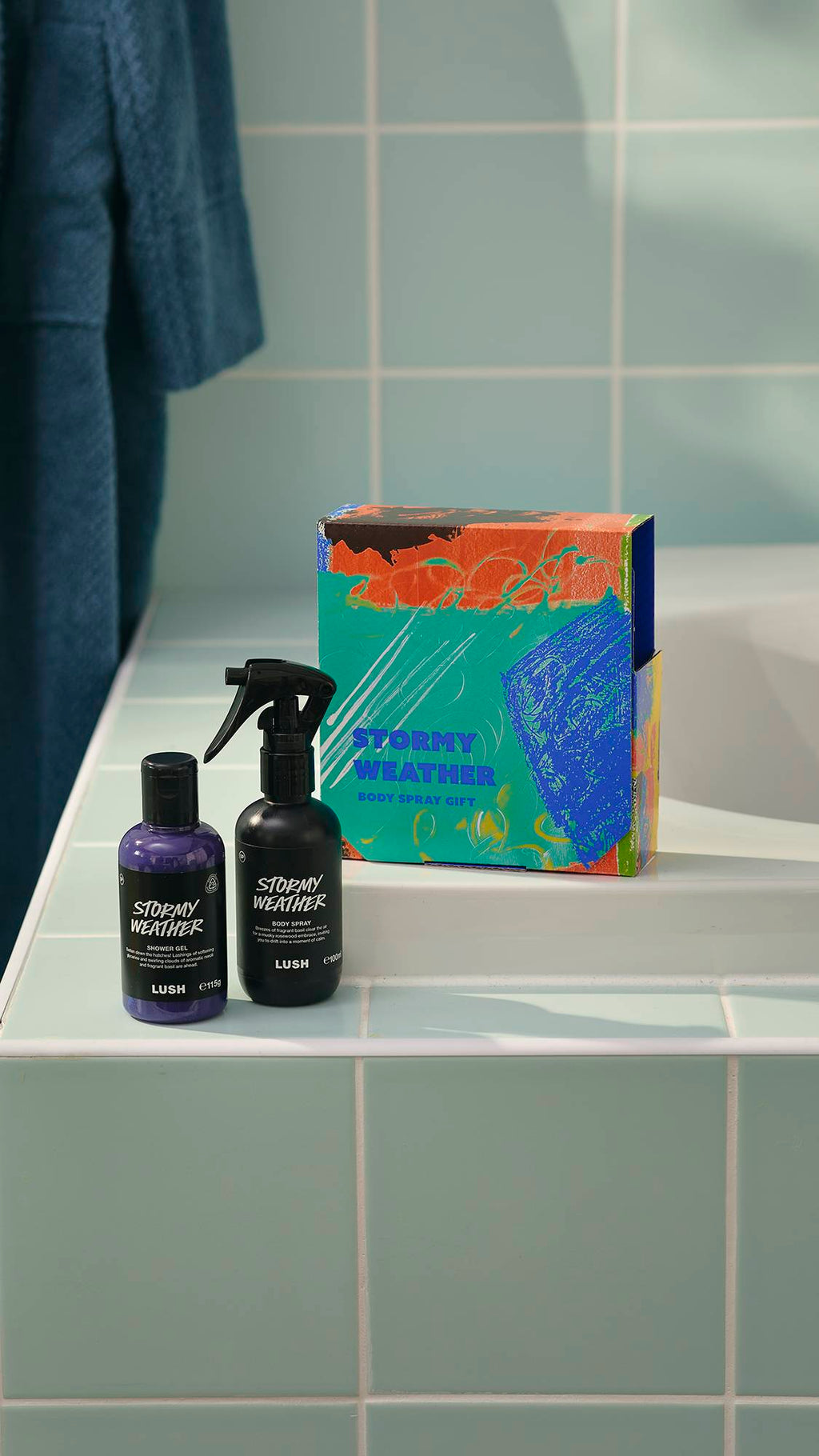 A Lush 'Stormy Weather' gift set sits on the edge of a bath against teal tiles. The set includes a colourful box, a bottle of purple shower gel, and a black bottle of body spray, both of the same name. A blue dressing gown hangs in the background.